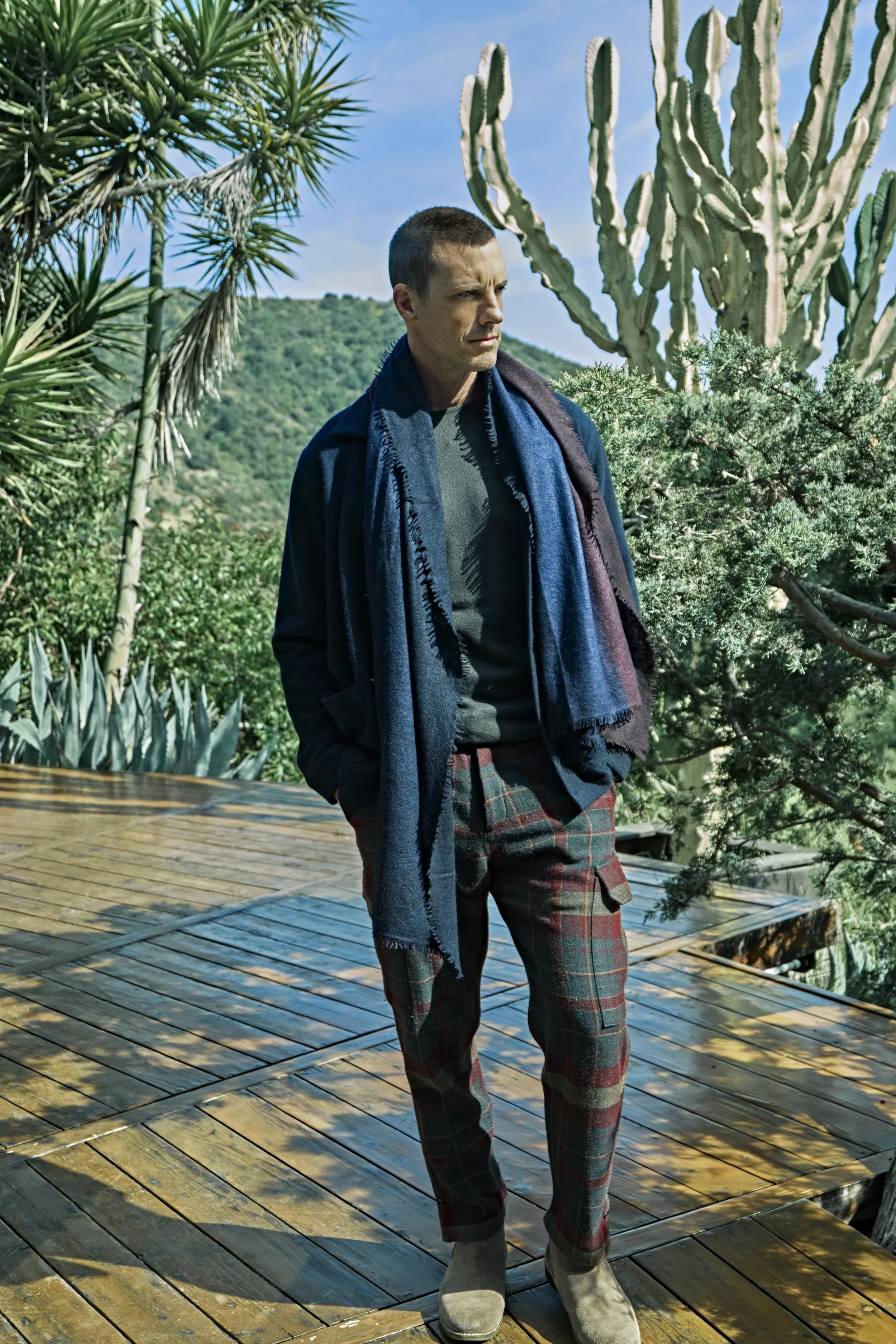 A man standing outdoors on a wooden deck, surrounded by desert plants including cacti and succulents, with a green hillside in the background.