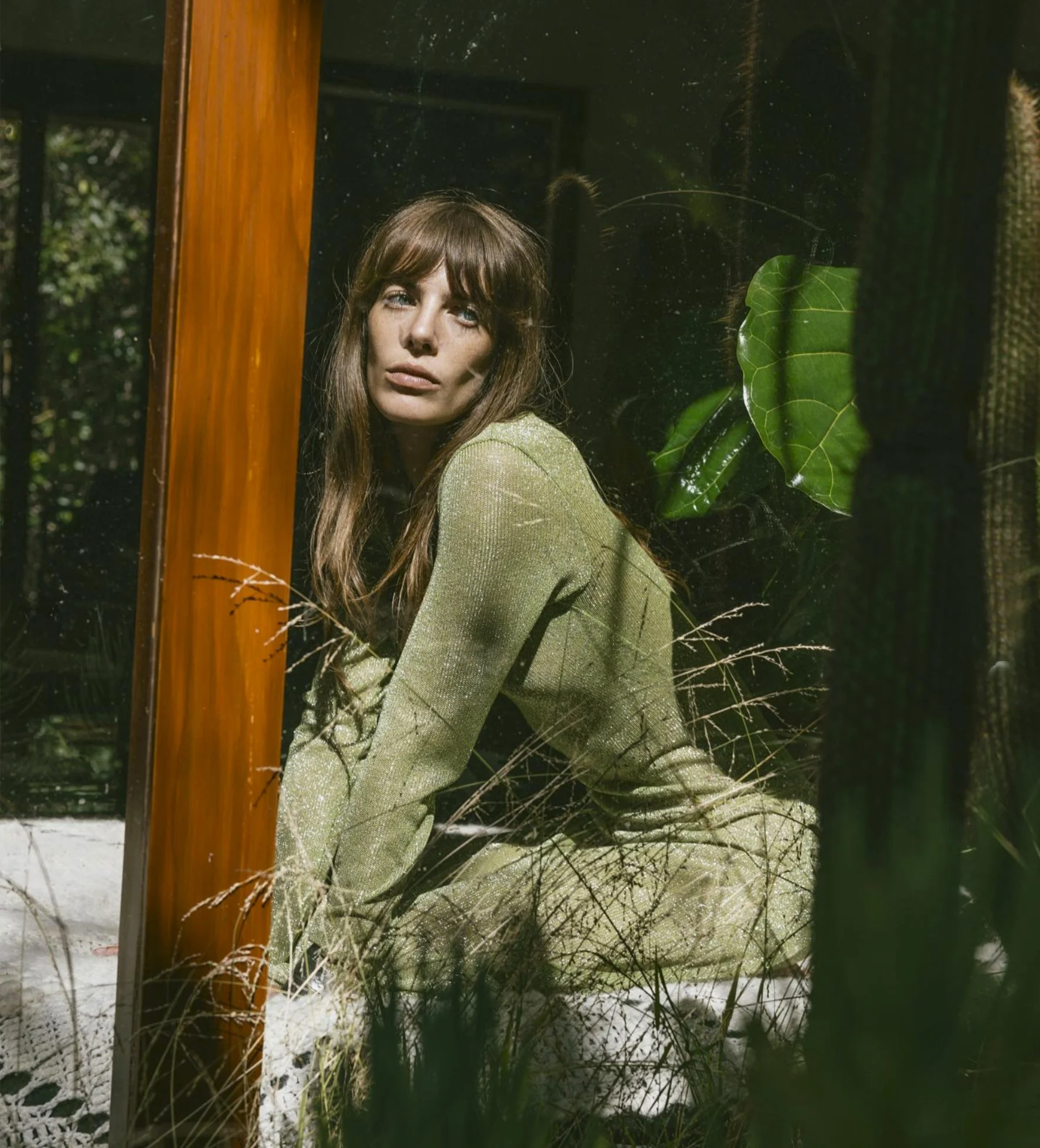 A woman with long brown hair in a green outfit sitting indoors, viewed through a window with reflections of plants and greenery outside.