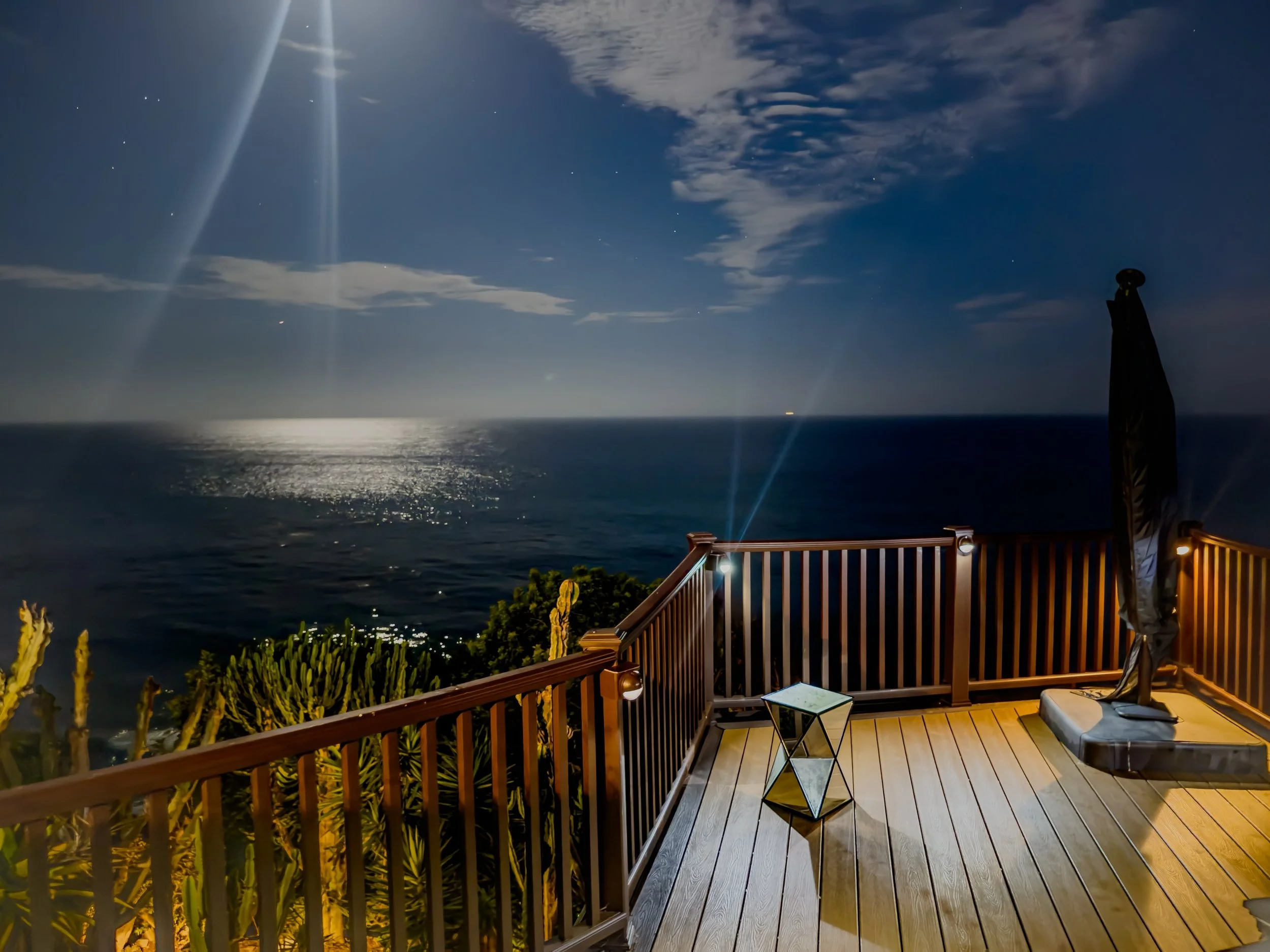 A nighttime ocean view from a wooden balcony with outdoor furniture, including a small table and chairs, overlooking the sea under a starry sky with clouds and a large moon.