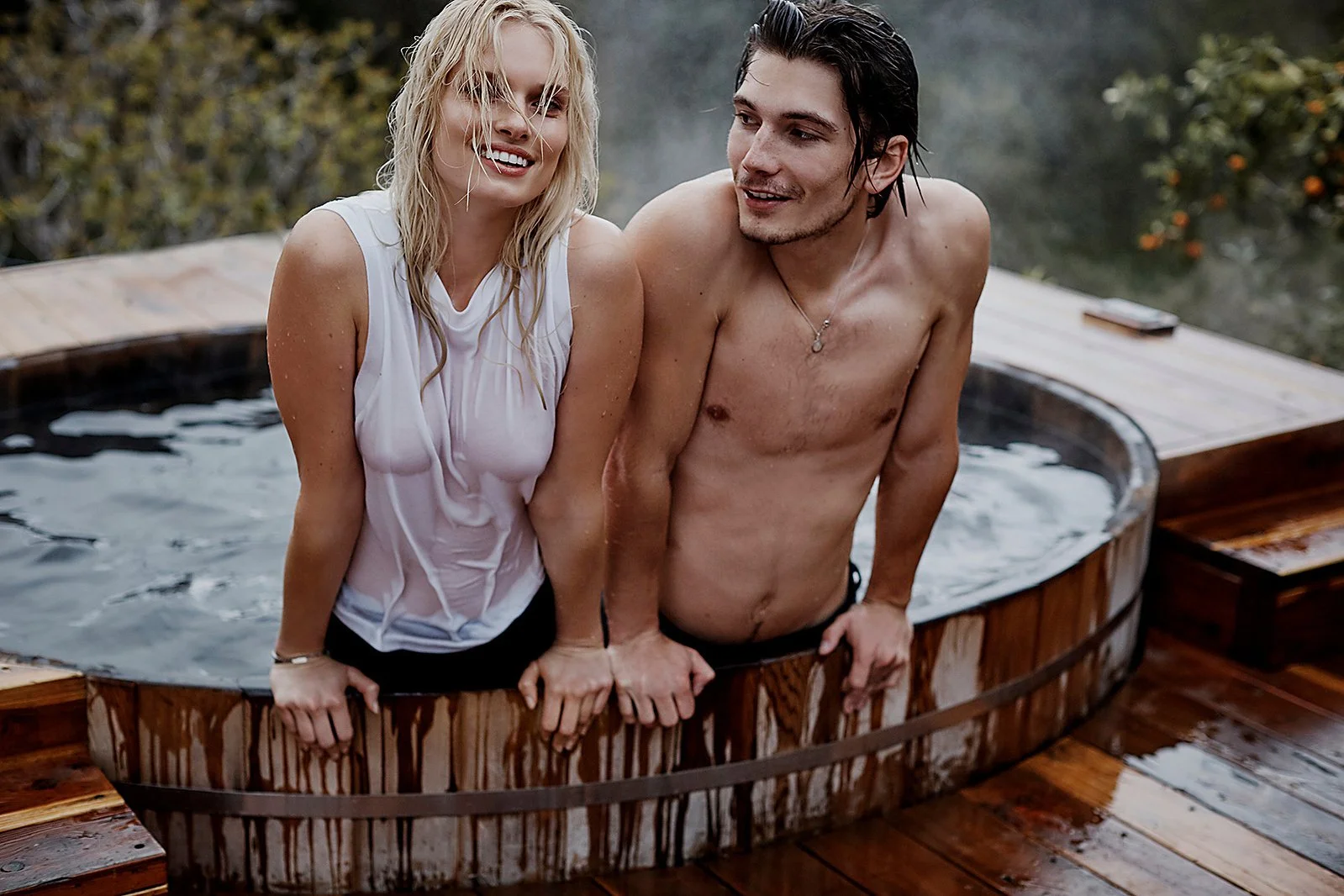 A smiling blonde woman and a shirtless dark-haired man sitting in a small wooden hot tub outdoors, holding hands and looking at each other.
