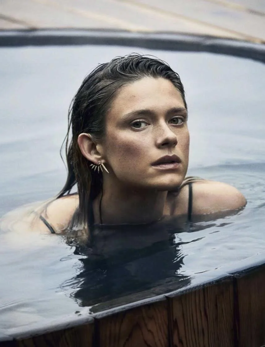 A woman with wet hair and earrings partially submerged in a hot tub or pool, looking at the camera.