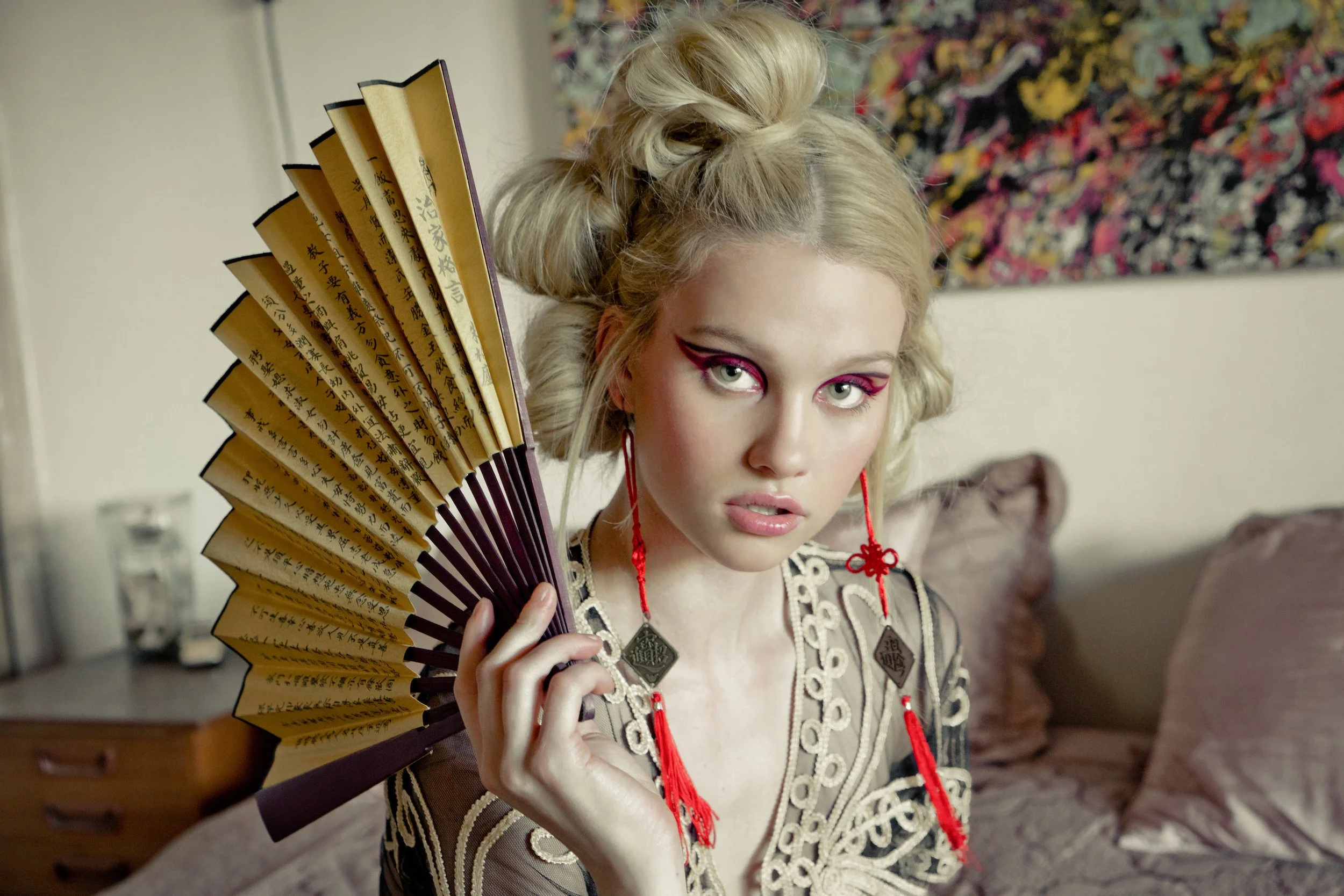A young woman with blonde hair styled in vintage curls holds an ornate folding fan with Japanese characters. She has dramatic eye makeup with pink eyeshadow and wears red earrings with intricate designs. The background features a colorful abstract pa
