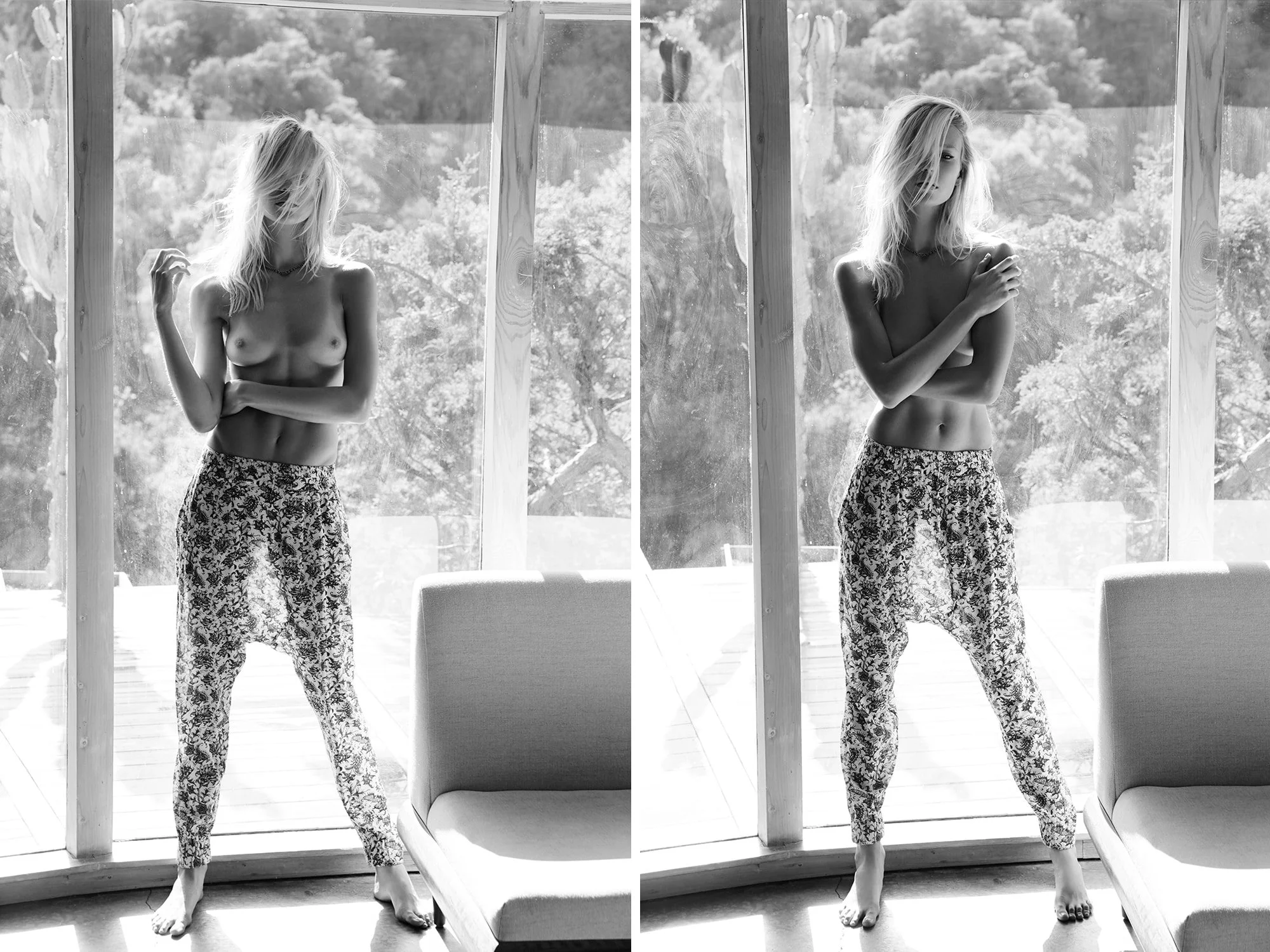 A black and white photograph of a woman standing by a large window with a view of trees outside. She is topless, wearing loose, patterned pants, barefoot, with long, wavy hair partially covering her face. Her pose varies in two different positions, o