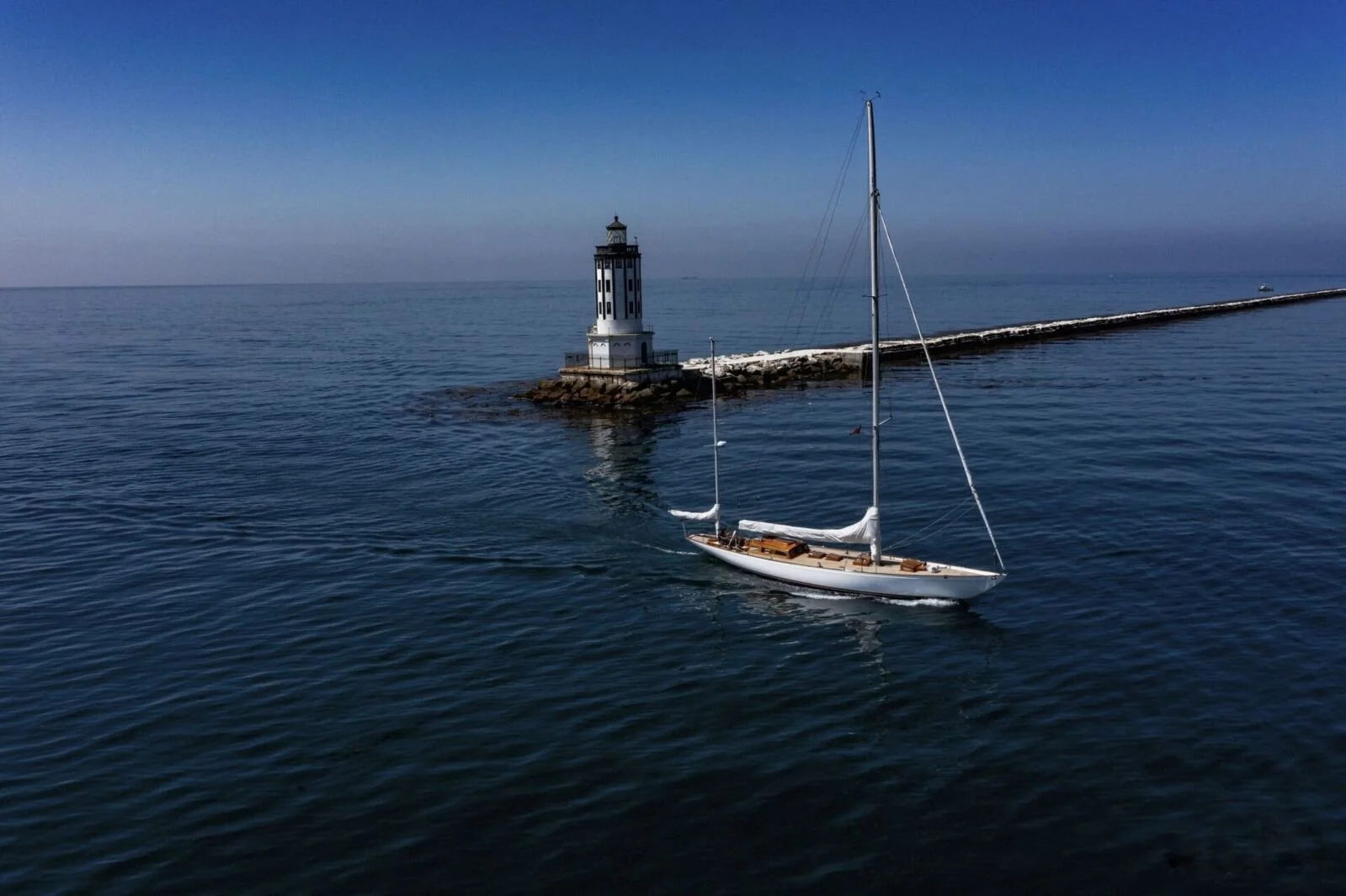 A sailboat in calm blue waters near a lighthouse on a breakwater.