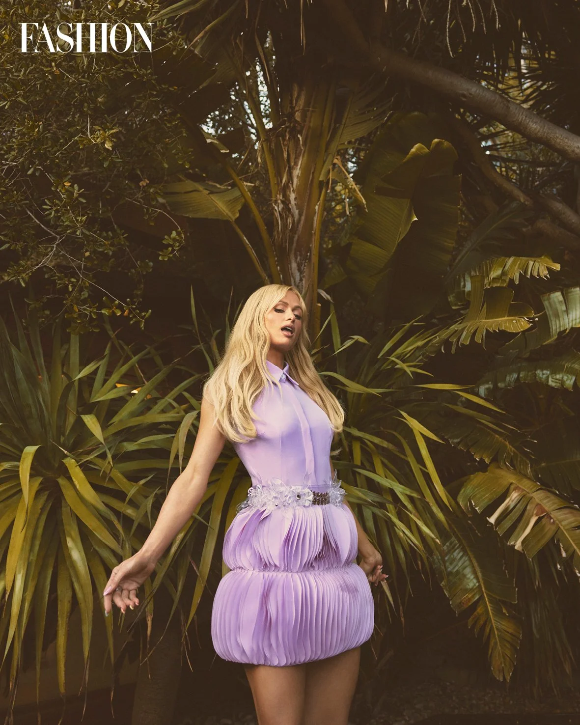 A woman with long blonde hair is standing in front of tropical plants, wearing a purple dress with a padded, pleated skirt and a decorative belt. The word 'FASHION' appears in the top left corner.