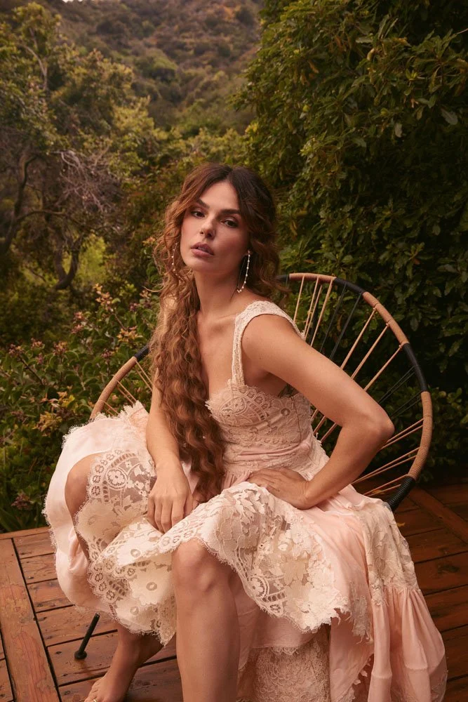 A woman with long, wavy brown hair, wearing a light pink lace dress and hoop earrings, sitting on a tan and black circular outdoor chair on a wooden deck surrounded by greenery and trees.