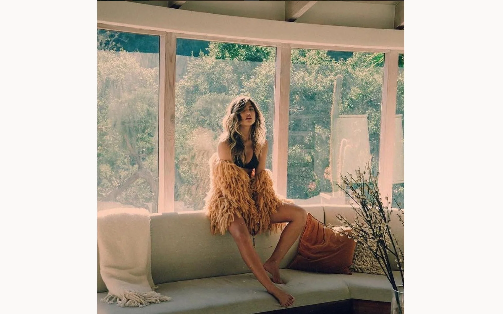 A woman with wavy hair sits on a beige sofa next to large windows, wearing a fluffy, tan faux fur coat and black lingerie, with sunlight streaming in and greenery visible outside.