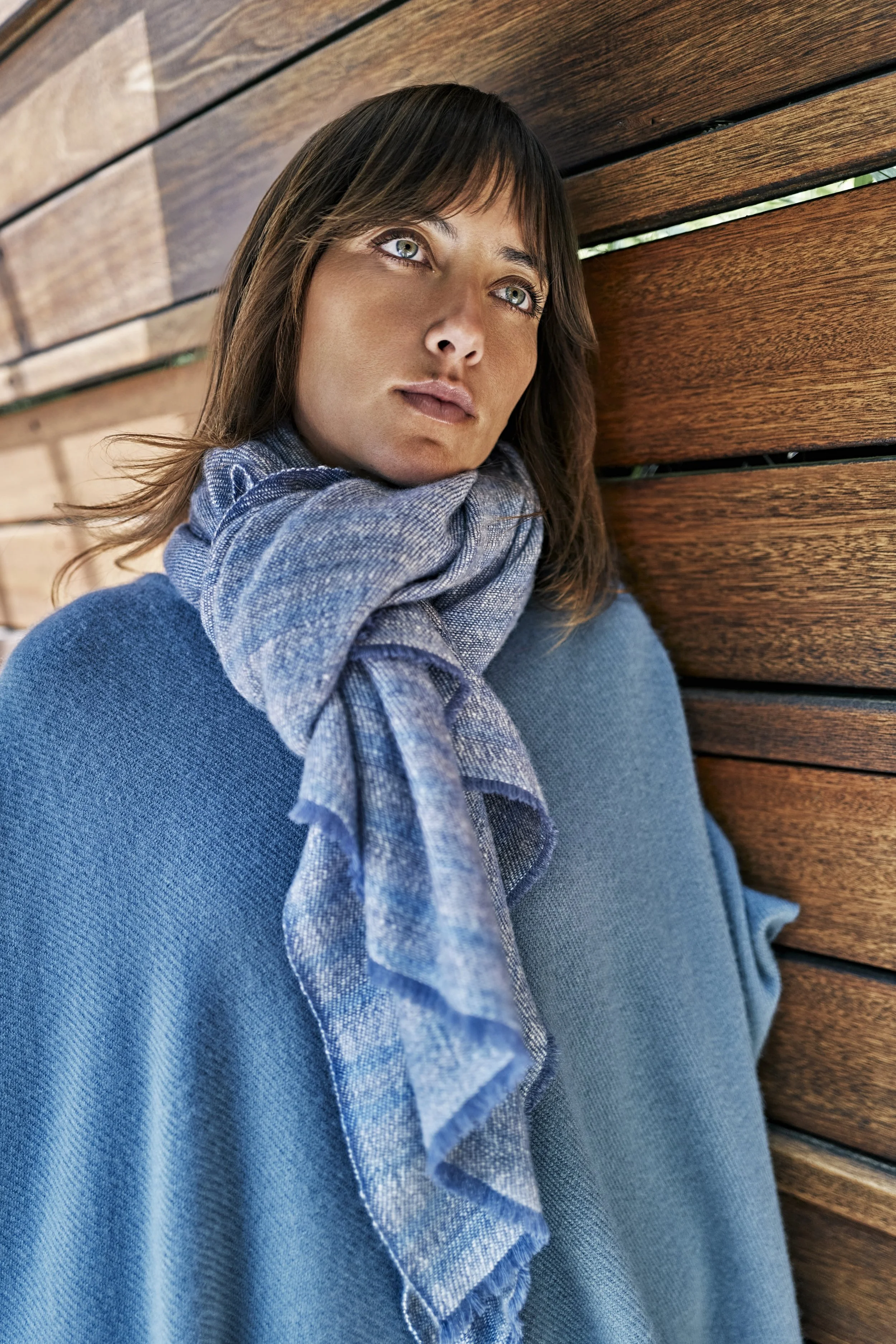 A woman with shoulder-length brown hair and blue eyes, resting her head against a wooden wall, wearing a blue sweater and a blue scarf.