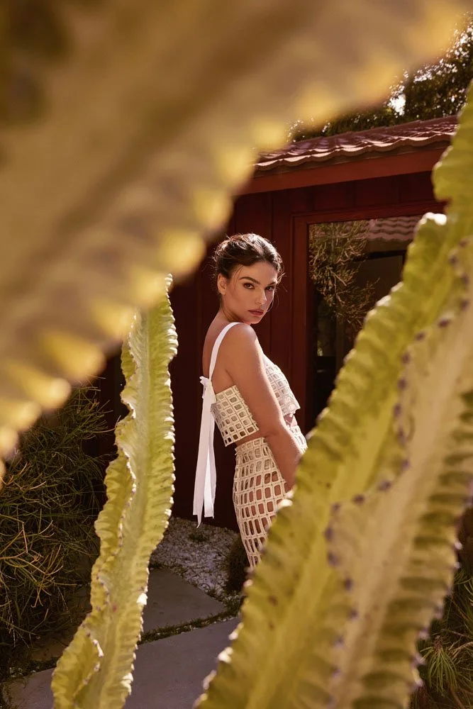 A woman with dark hair in a bun stands outdoors behind large, green, tall, leafy plants, looking at the camera with a serious expression.