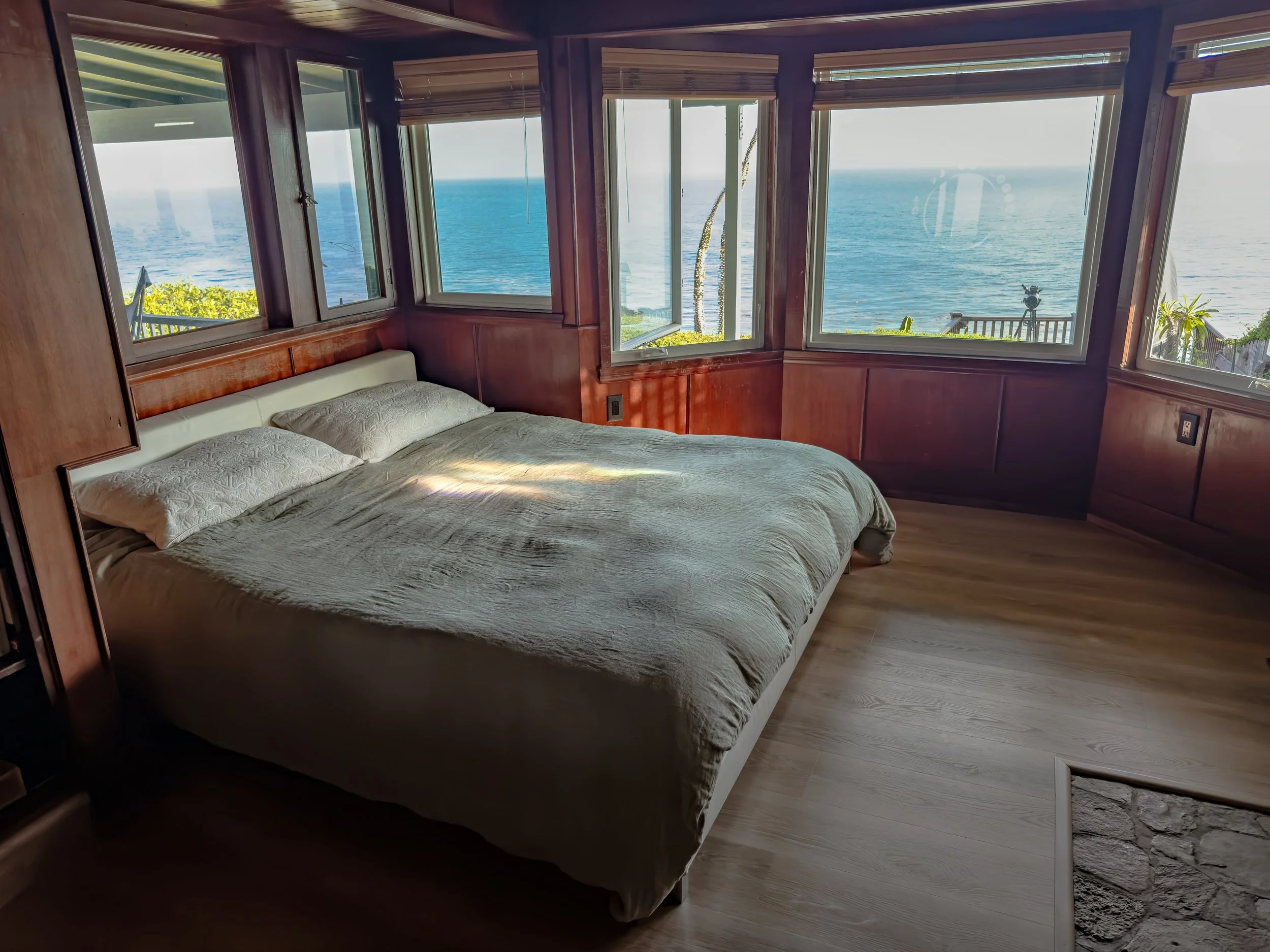A bedroom with wooden walls and a bed with beige bedding, large windows offering a view of the ocean and sky, and a tropical outdoor scene with greenery and a deck visible outside.