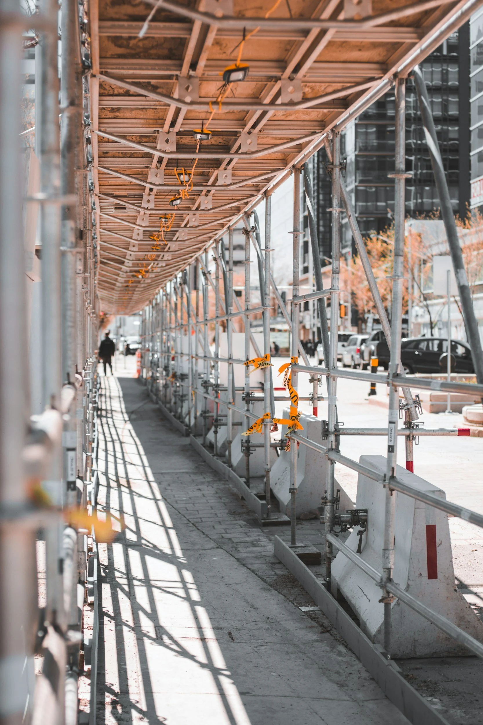 Construction scaffolding and safety barriers along a city sidewalk, with orange warning tape and shadow patterns cast on the ground.