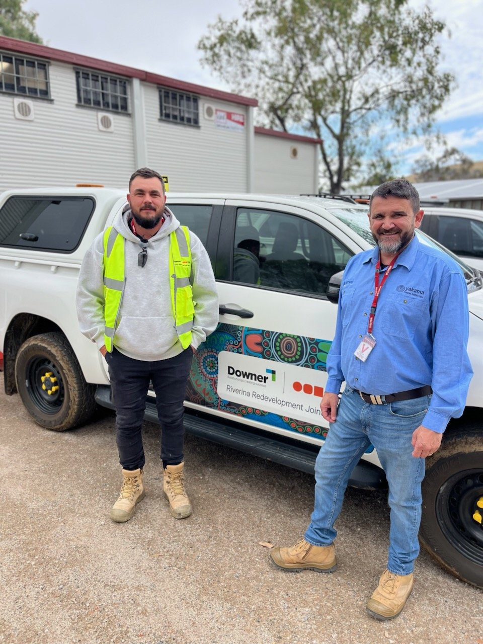 Two men standing outdoors next to a vehicle with a logo for Downer and Riverina Redevelopment Joint Venture, smiling at the camera. One man is wearing a yellow safety vest and hoodie, and the other is wearing a blue shirt with a badge. The vehicle is white with decorative panels, and background includes buildings and trees.