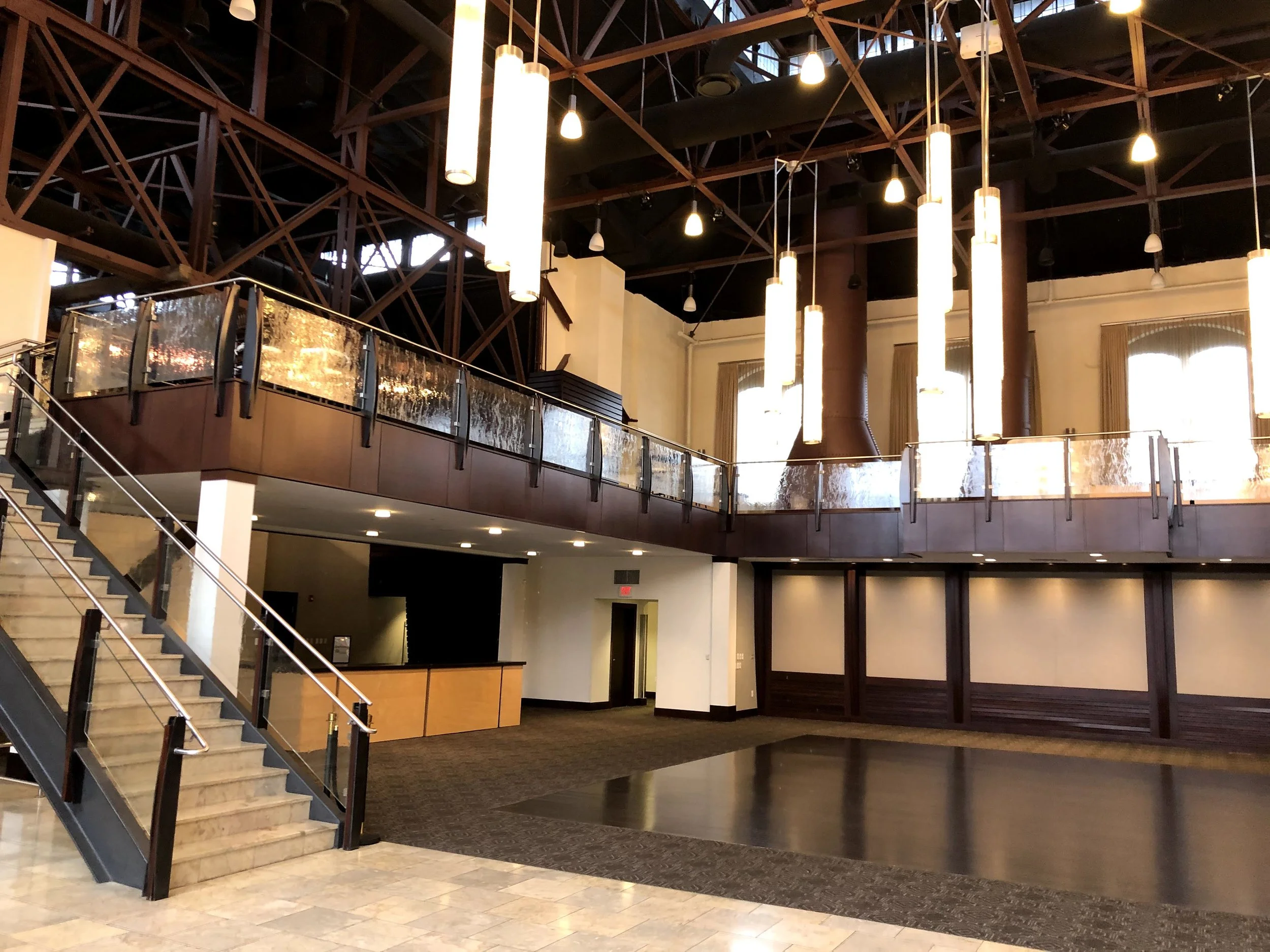 Interior of a spacious, modern building with high ceilings, hanging cylindrical lights, a staircase with glass and metal railing, and a balcony overlooking the main floor.