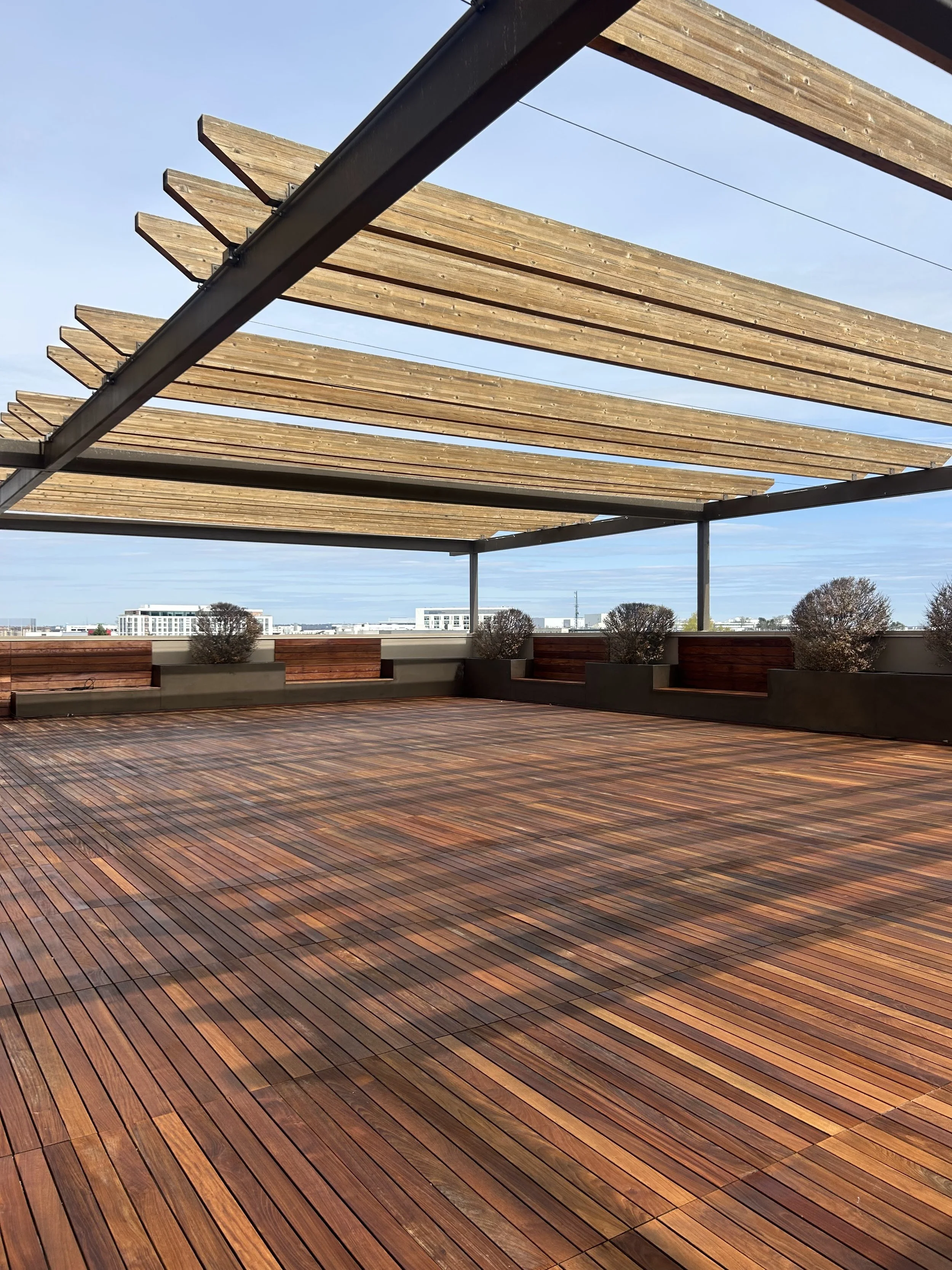 Rooftop terrace with a wooden floor and a pergola above, with planters and trees along the perimeter and cityscape in the distance.