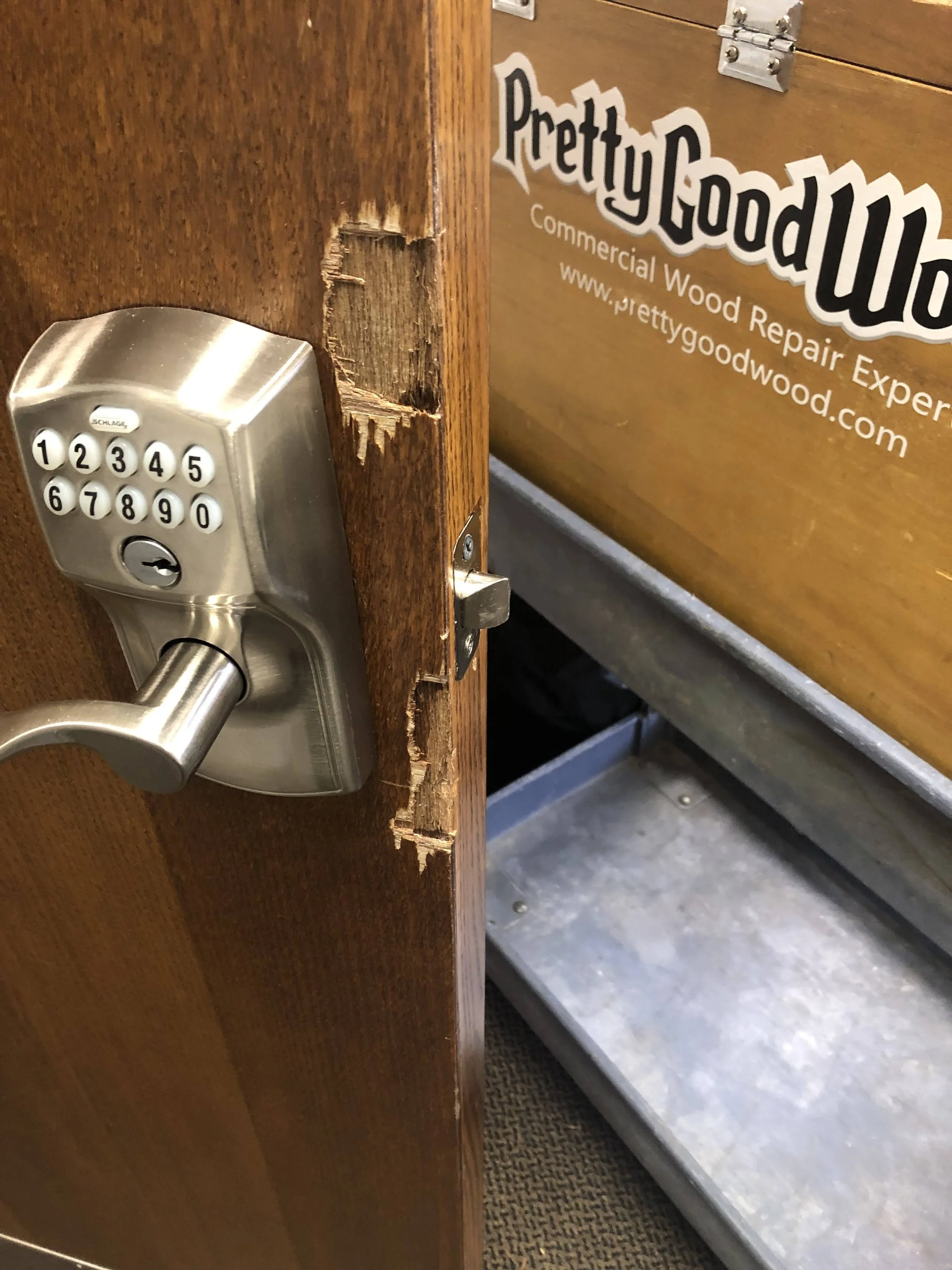 A wooden door with a keypad lock and a hole around the latch area, revealing damage to the wood. In the background, there is a sign for Pretty Good Wood, a commercial wood repair business.