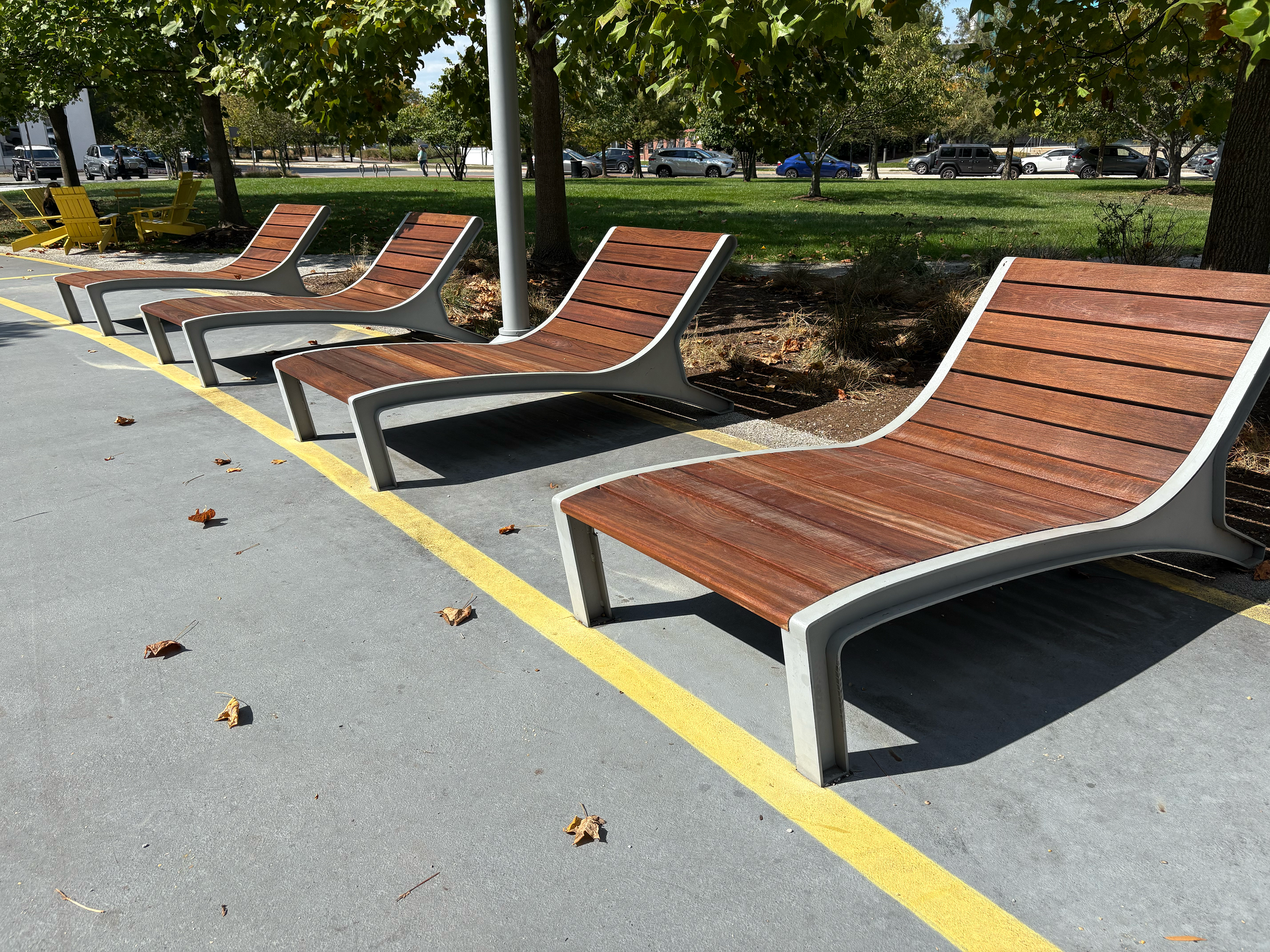 Four empty wooden and metal park benches are arranged in a row along a concrete sidewalk, with trees and parked cars in the background on a sunny day.
