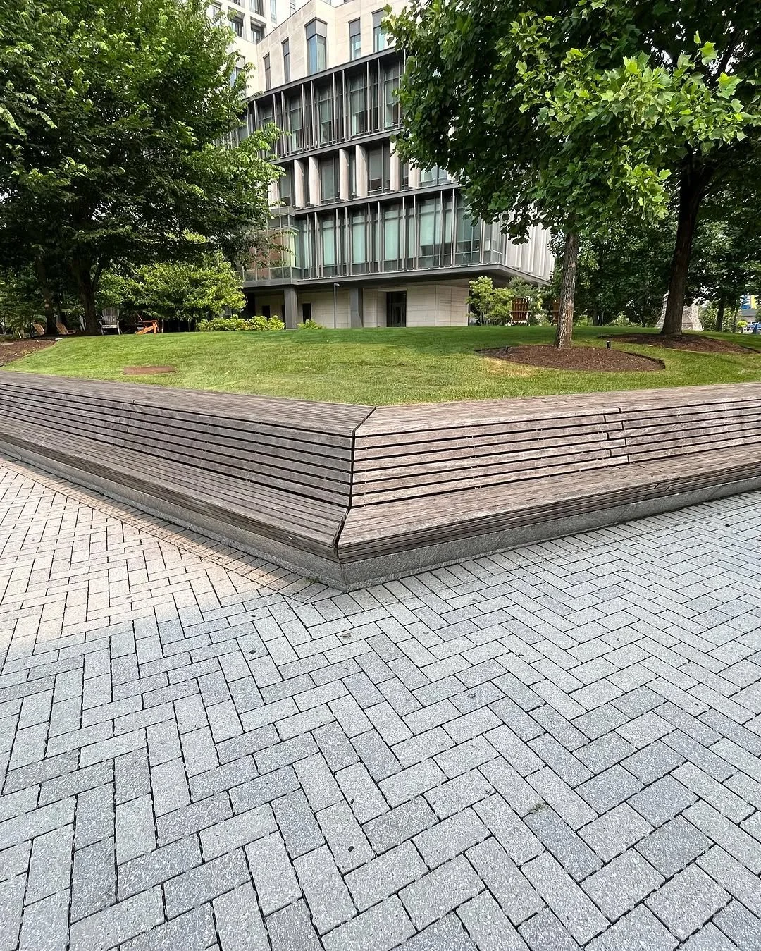 A landscaped urban park area with a curved wooden bench, green grass, trees, and a modern multi-story building in the background.