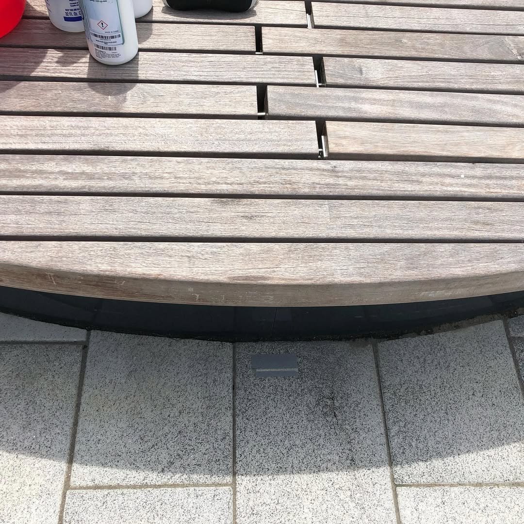 A wooden outdoor table with visible gaps between the planks, partially shaded on a tiled surface, with bottles placed on the top.
