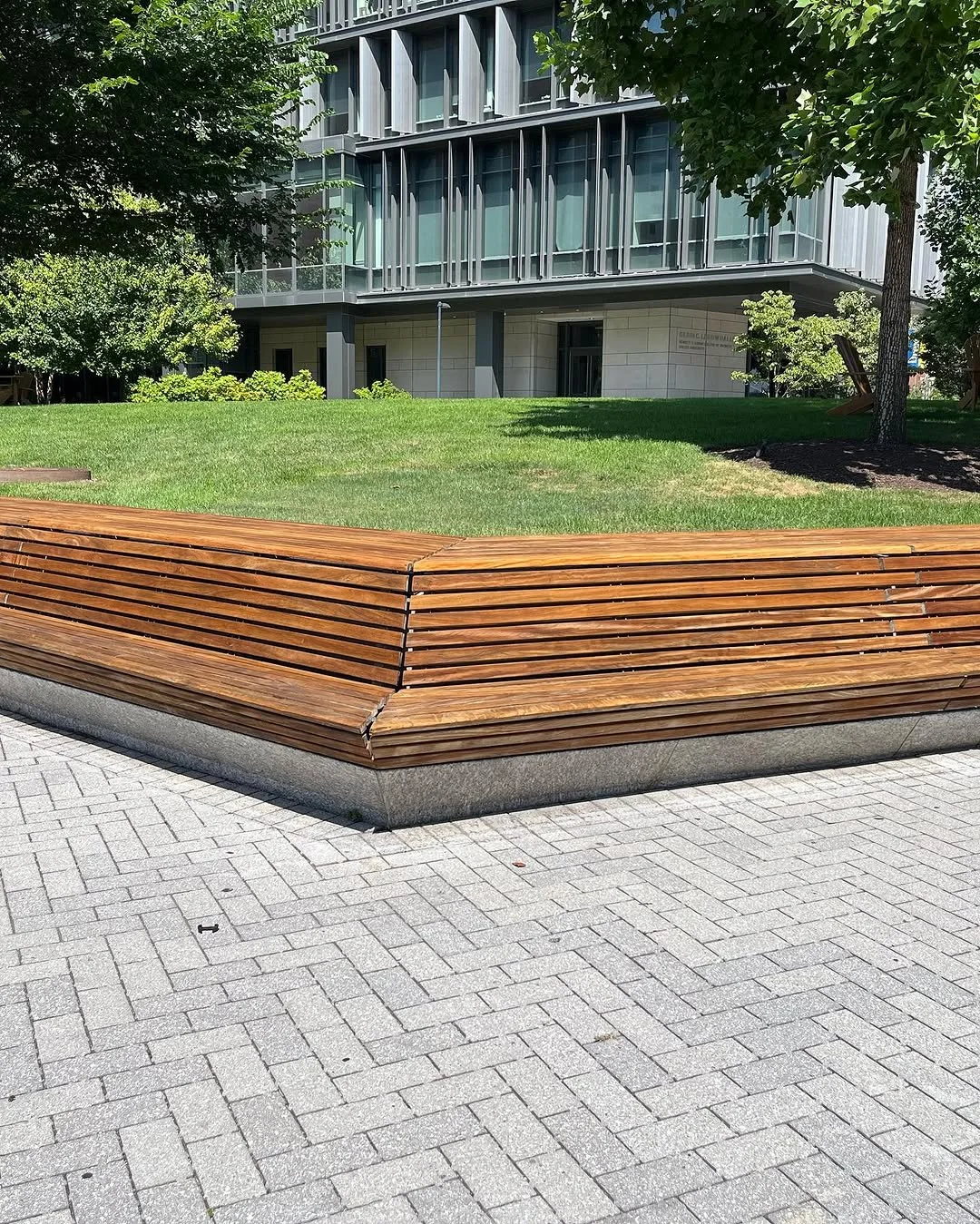 A modern urban park with a curved wooden bench on a brick-paved surface, green grass, trees, and a multi-story building with glass windows in the background.