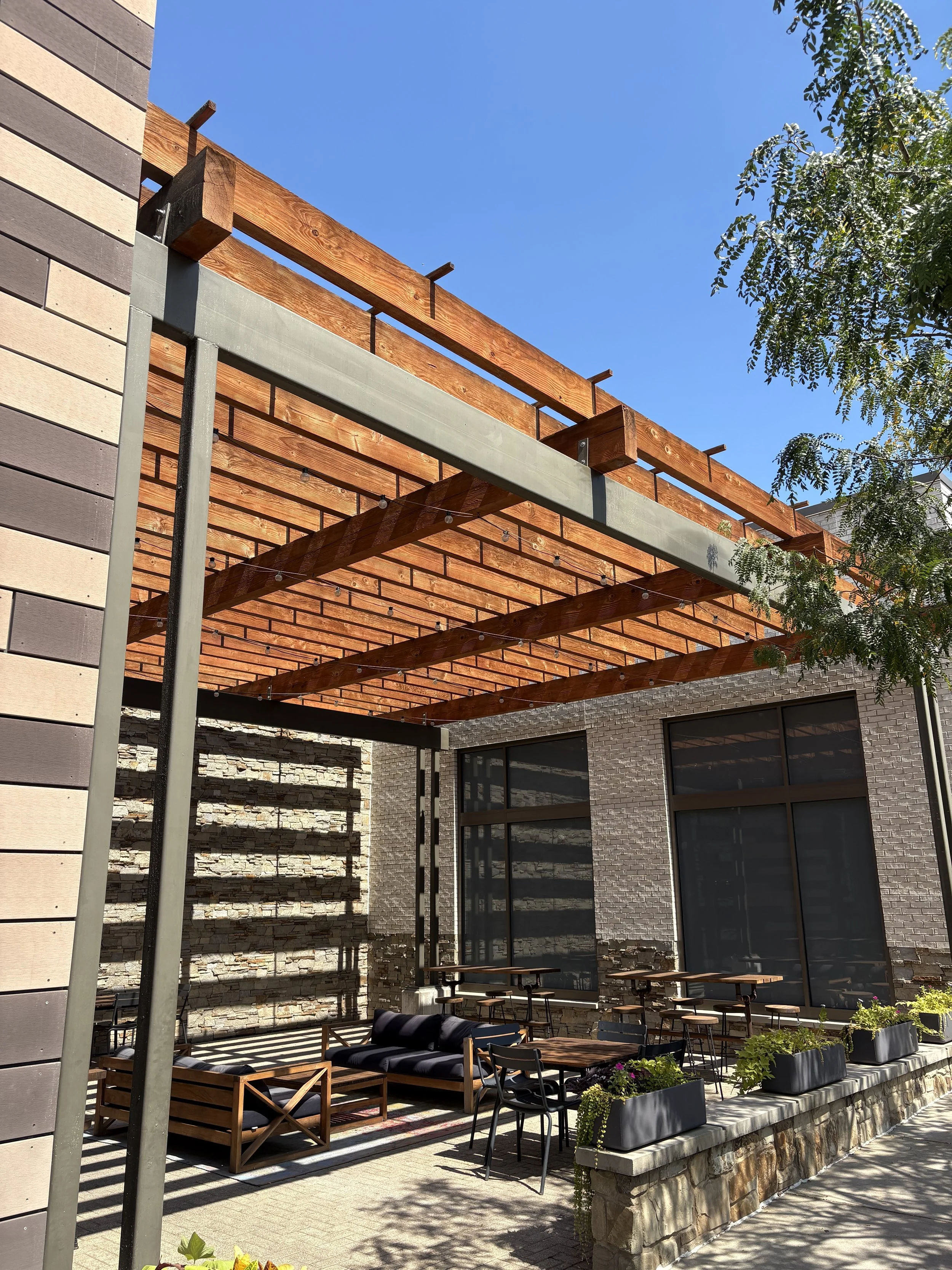 Outdoor patio area with wooden pergola, seating including a black striped sofa and chairs, potted plants, stone wall, and large windows.