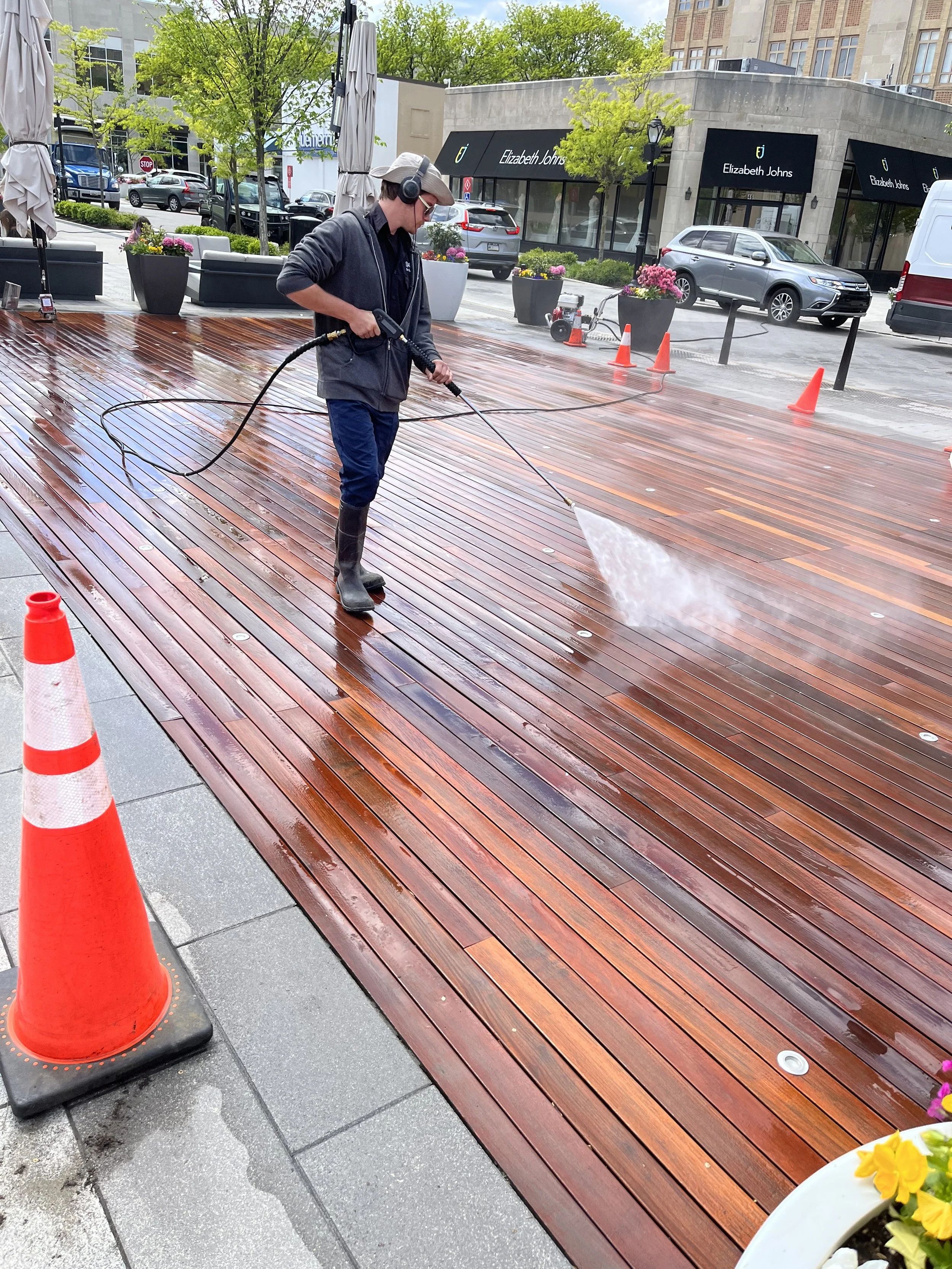 A worker power-washing a wooden outdoor deck with orange safety cones nearby, in an urban outdoor setting.