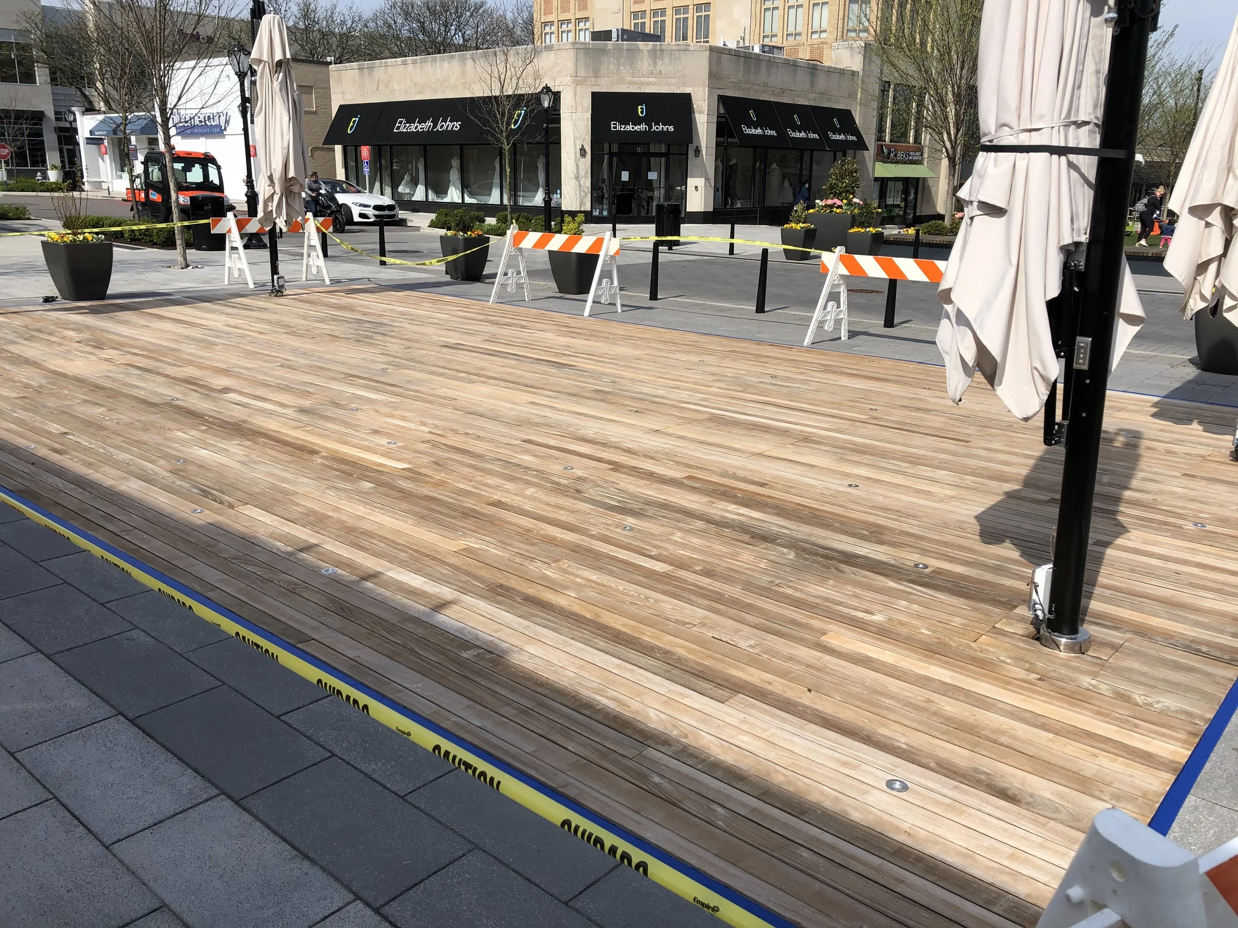 Empty outdoor stage with wooden flooring surrounded by caution tape and barricades, with closed umbrellas on either side, in a shopping district with storefronts and parked vehicles in the background.