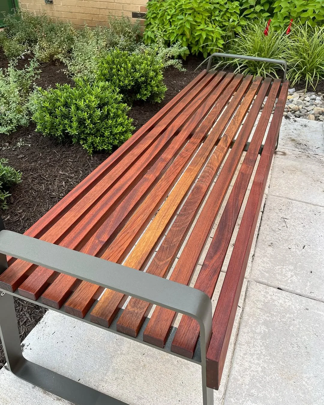 Wooden park bench with gray metal armrests on a concrete sidewalk, surrounded by green bushes and plants in a garden setting.