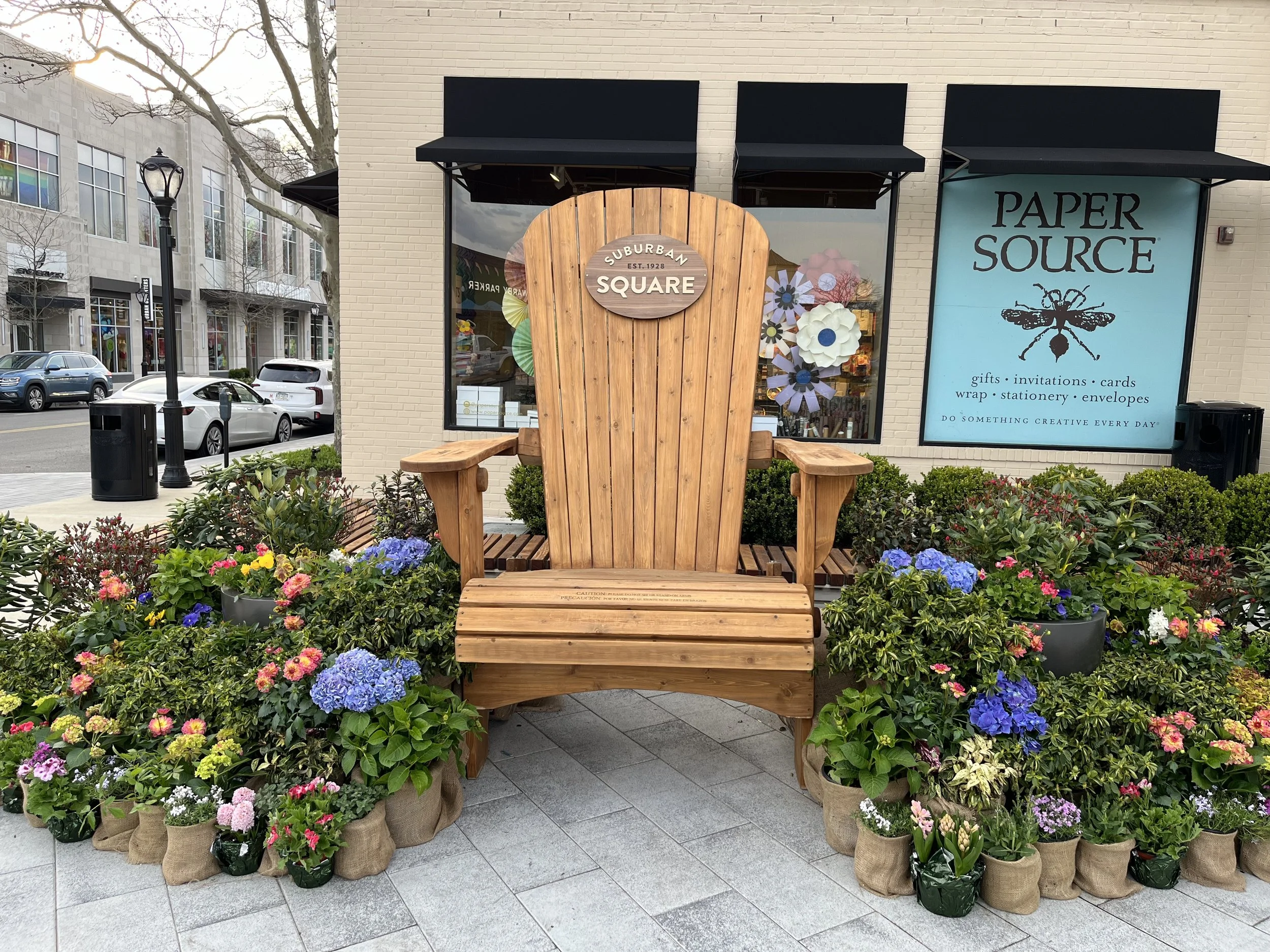 A large wooden Adirondack chair with a circular sign on top reading "Suburban Square" is surrounded by colorful flowers in pots along the sidewalk outside a building with signs for Paper Source and other stores.