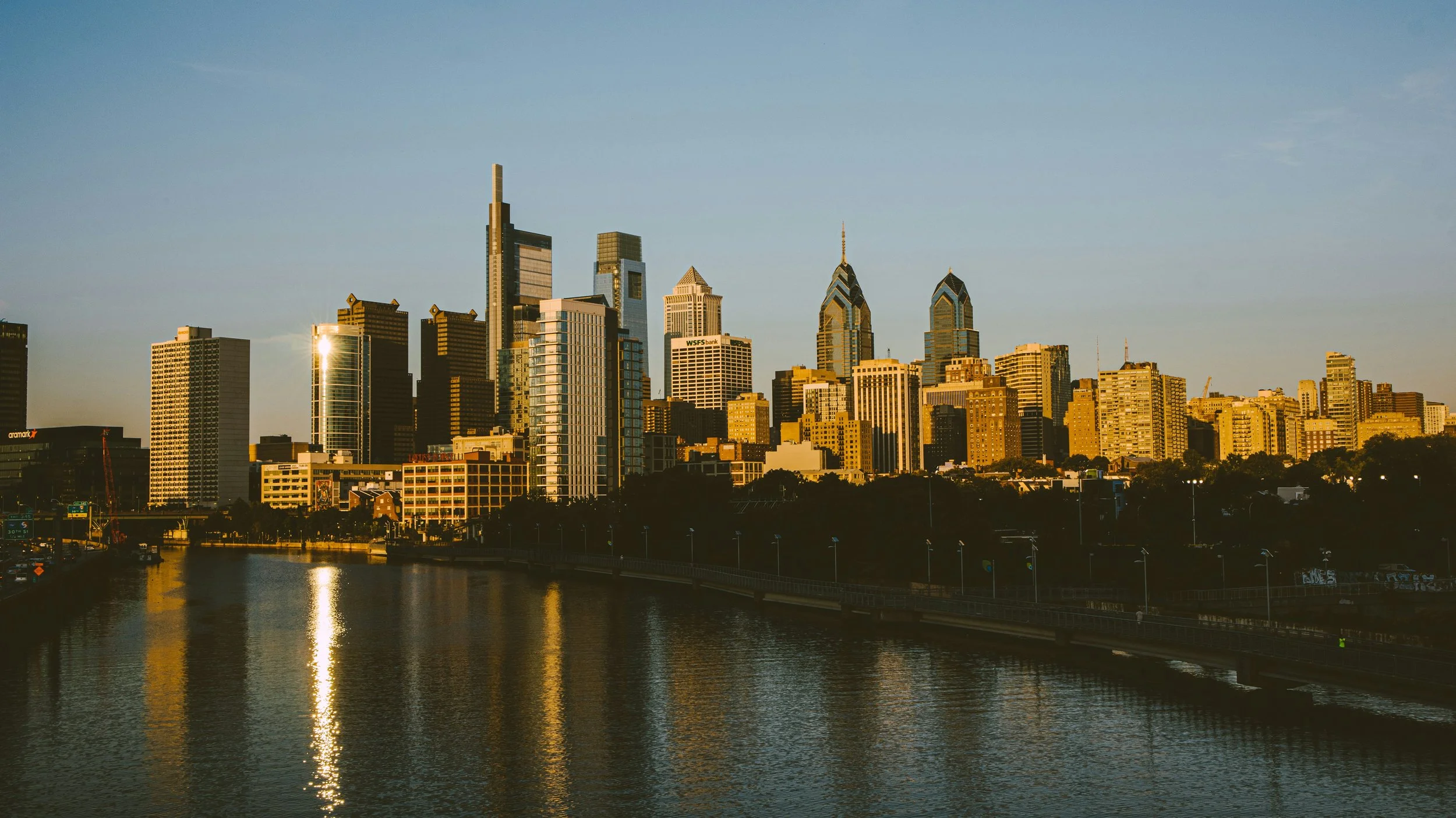 Sunset city skyline with tall skyscrapers, a river in the foreground reflecting the buildings, and a clear sky.