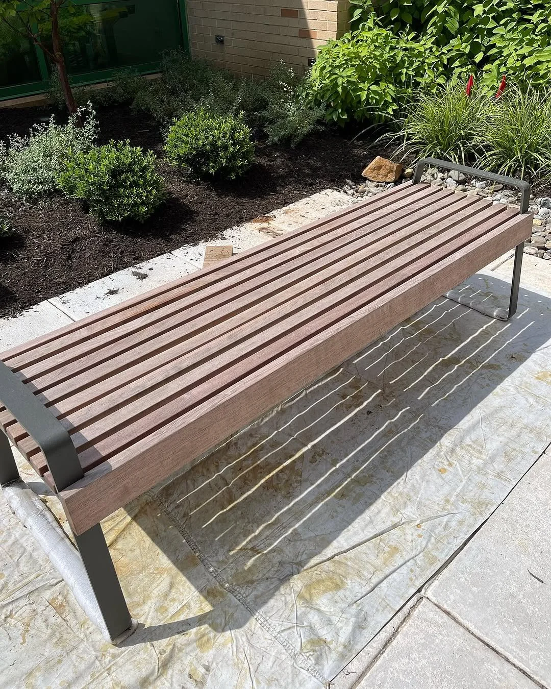 A wooden outdoor bench with metal legs placed on a fabric outdoor cover, surrounded by a garden with bushes and plants, against a brick wall.
