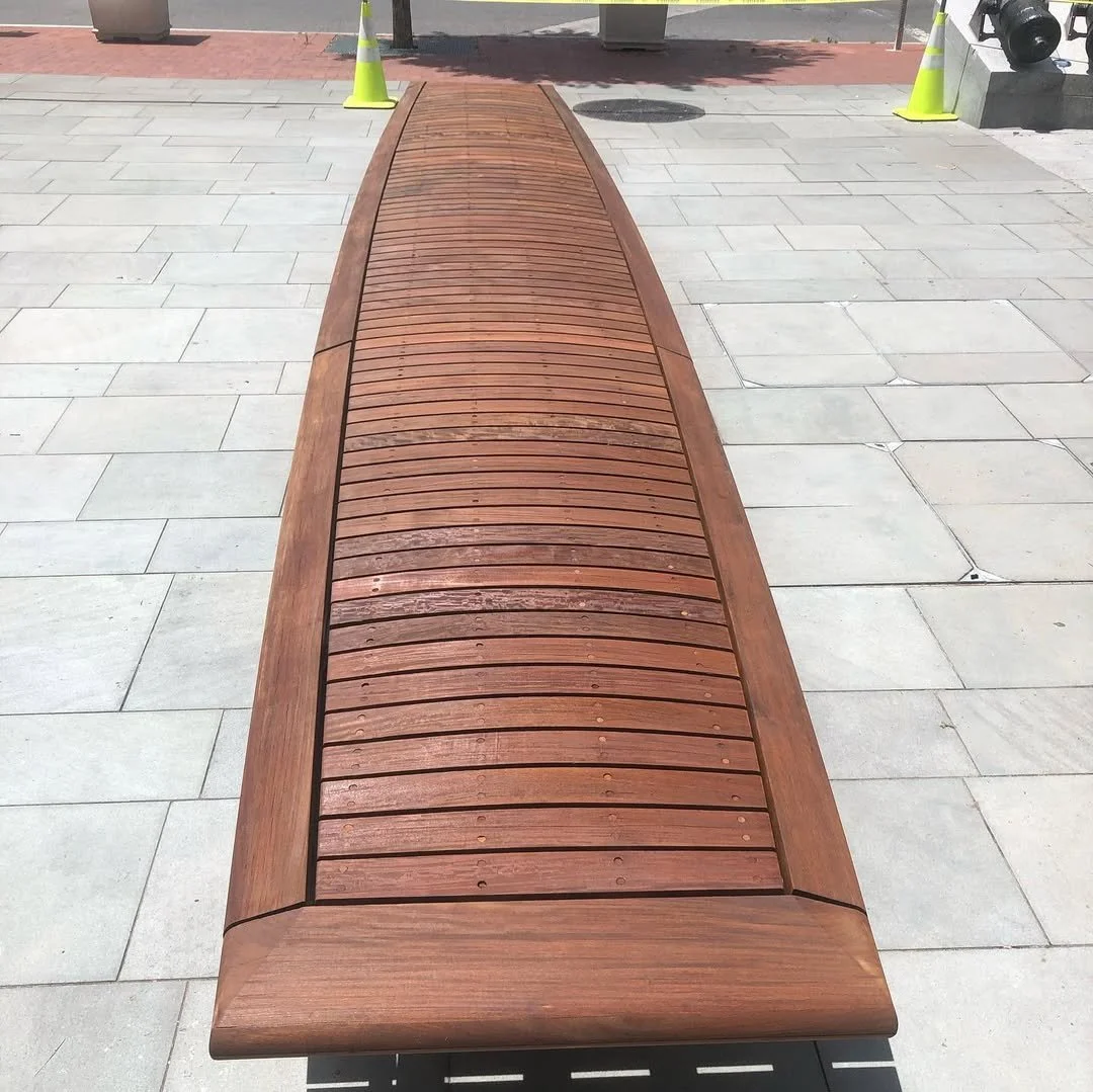 A wooden bench on a paved walkway with yellow traffic cones and potted plants in the background.