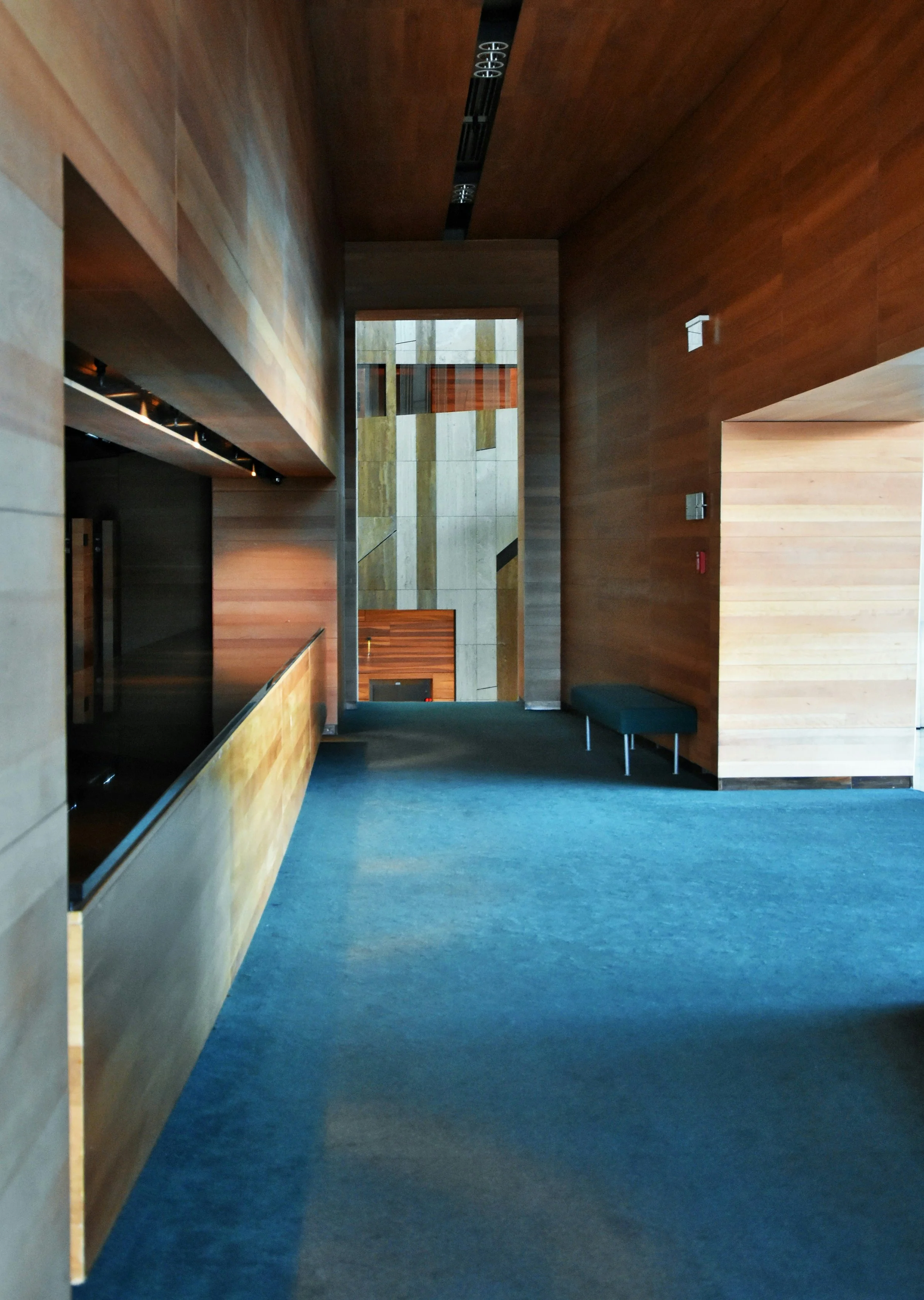 Interior view of a building with wooden walls, carpeted floor, and a bench near the wall, showing corridor leading to a less illuminated area with a combination of wood and stone walls.