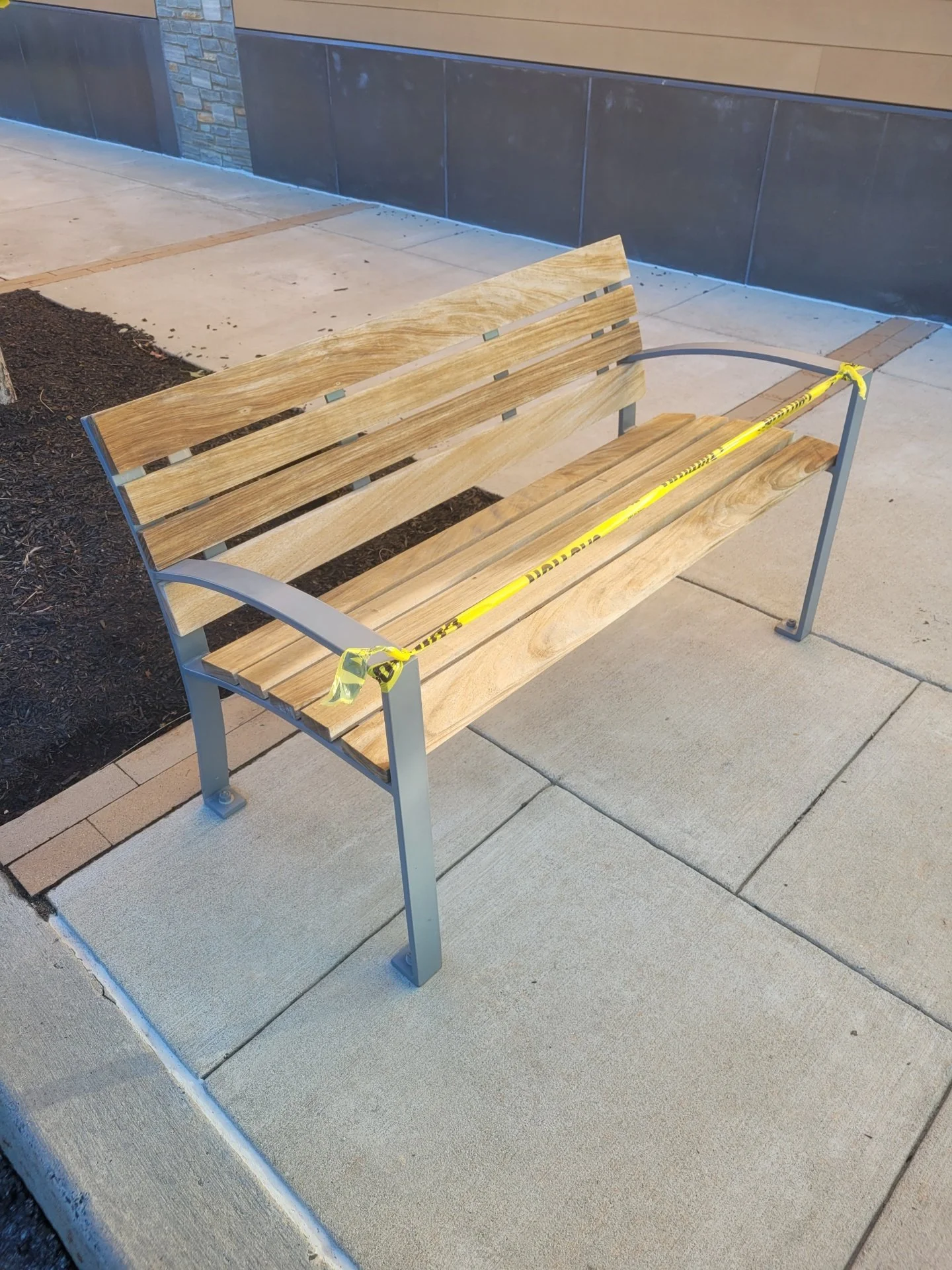 Wooden park bench with gray metal frame, cordoned off with yellow caution tape on a sidewalk.