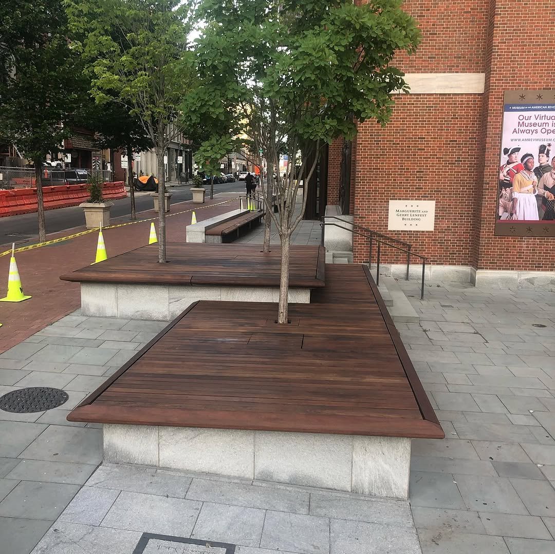 Street view with wooden benches surrounding trees, brick building, sidewalk, and poster on the building.