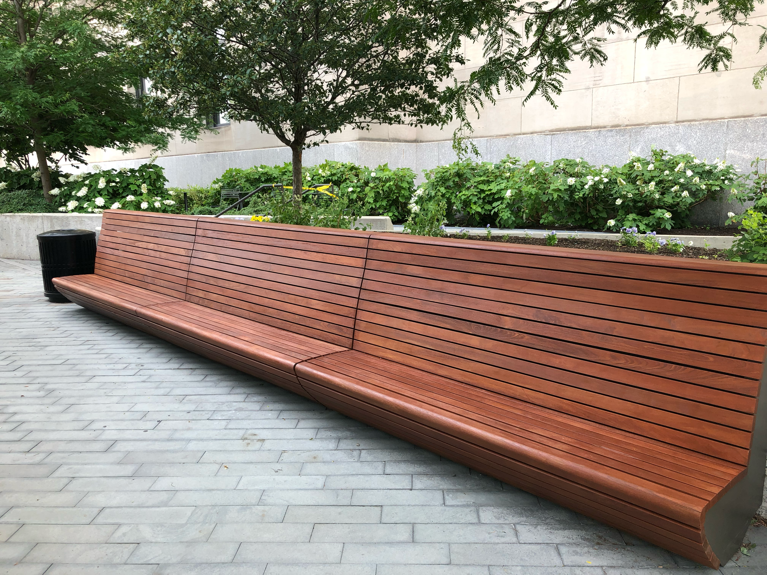 A long empty wooden park bench with slats, situated on a paved area with a black trash can nearby, surrounded by greenery and trees.
