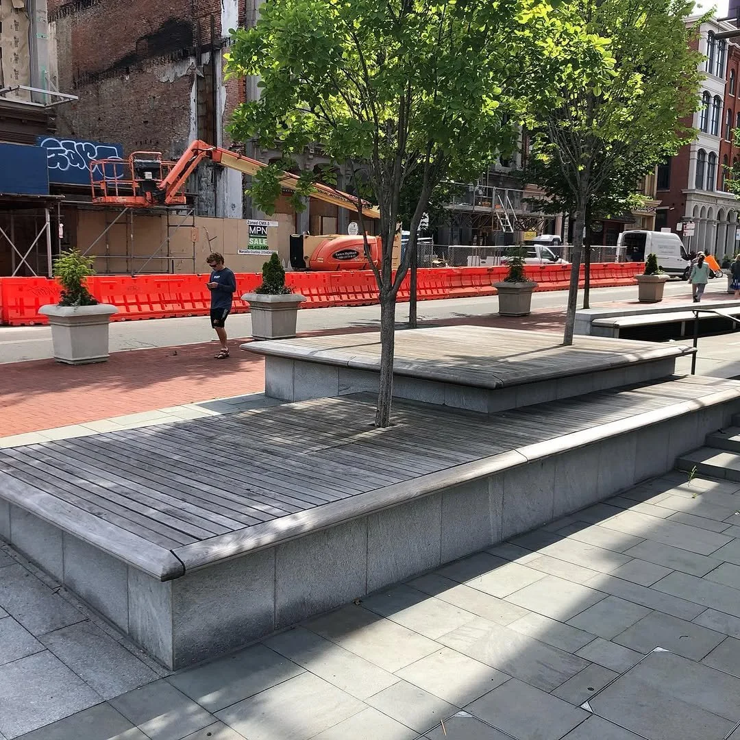 City street scene with trees, benches, and people walking, with construction work visible in the background.