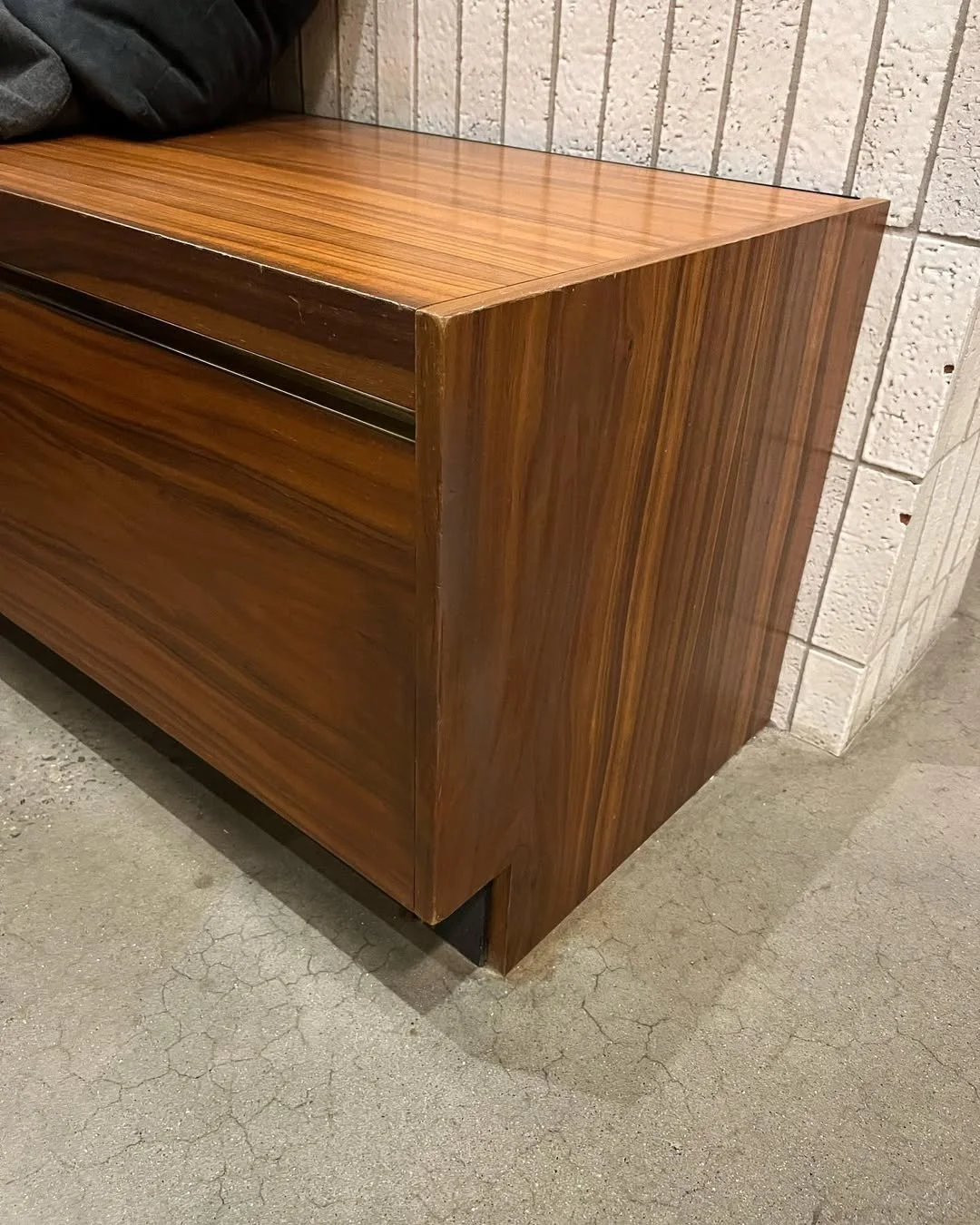 A wooden cabinet with a smooth, polished surface, positioned against a textured concrete block wall, standing on a cracked concrete floor.