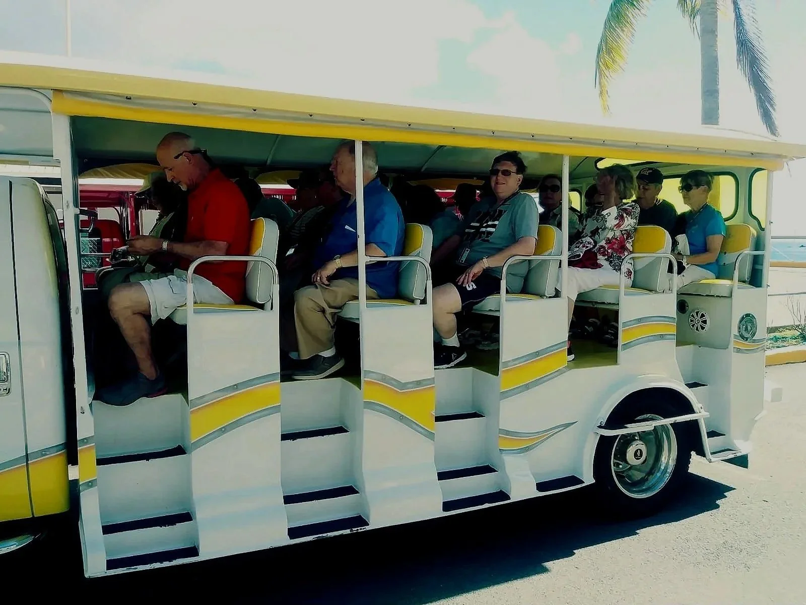 People sitting on a yellow and white open-air tram with a yellow canopy, traveling along a sunny street with palm trees in the background.