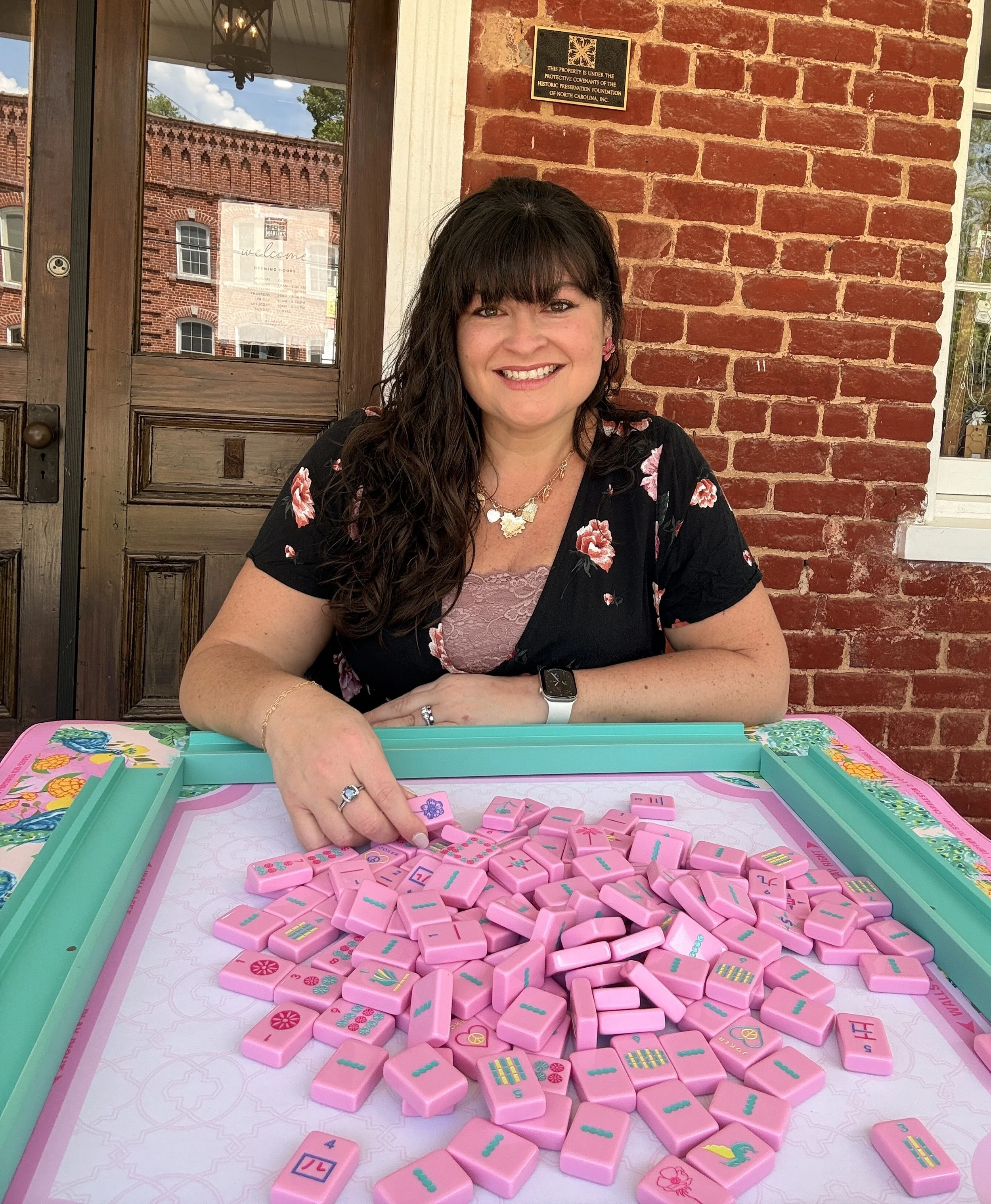 A woman with long dark hair, bangs, and pink earrings seated at a table playing with pink Mahjong tiles featuring flowers, animals, and symbols. She is smiling, wearing a dark floral top, a gold necklace, and an Apple Watch. The setting is a brick-walled restaurant or café with a wooden door, brick walls, and large windows, one displaying a sign with hours of operation.