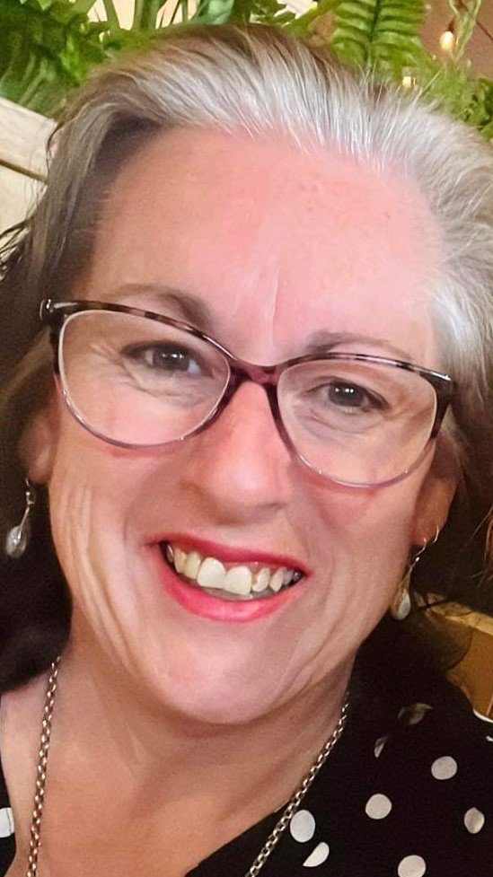 Close-up photo of a smiling woman with glasses, earrings, and pearls, with greenery in the background.