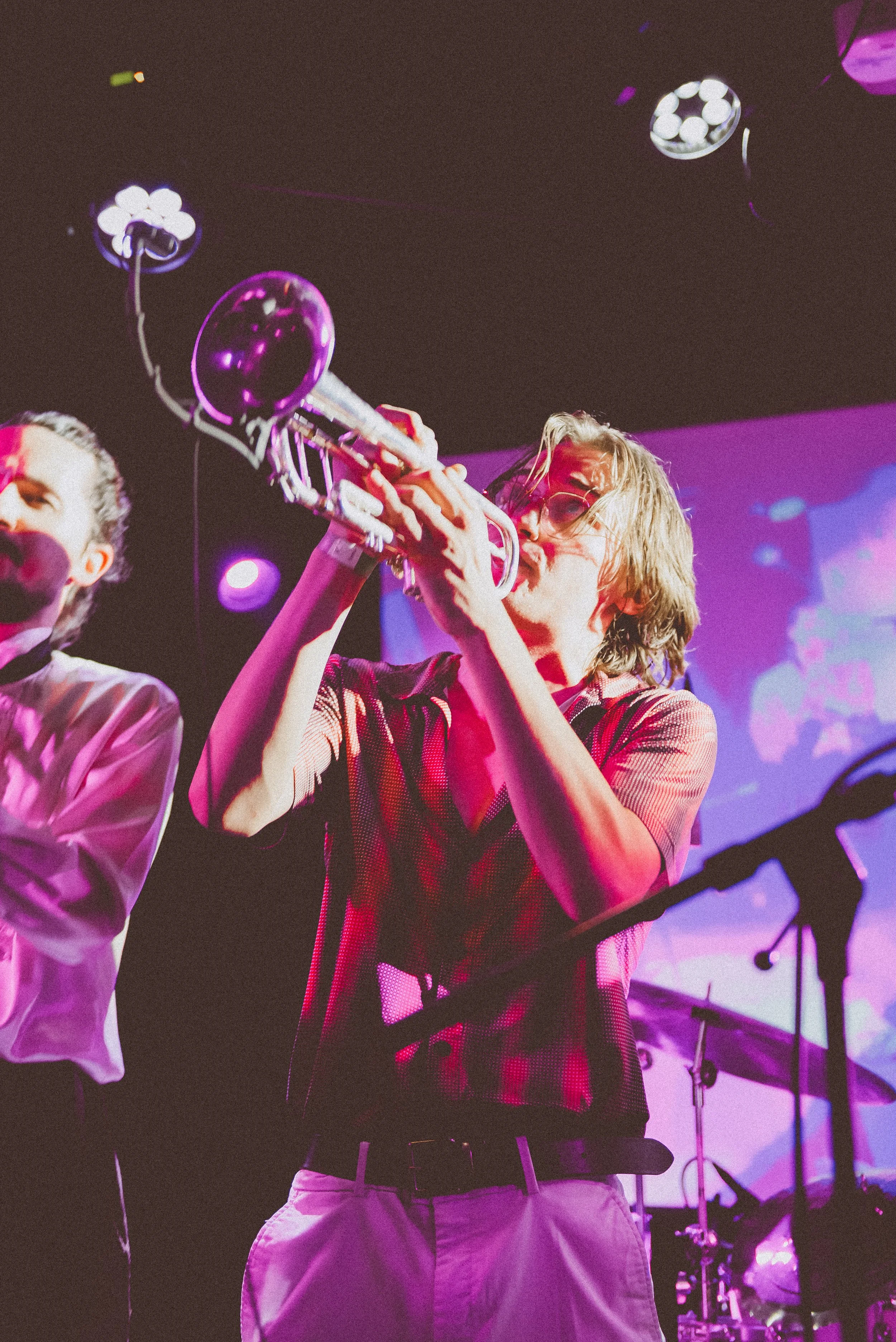 Musician with shoulder-length hair and glasses playing a trumpet on stage with purple lighting and a drum set in the background.