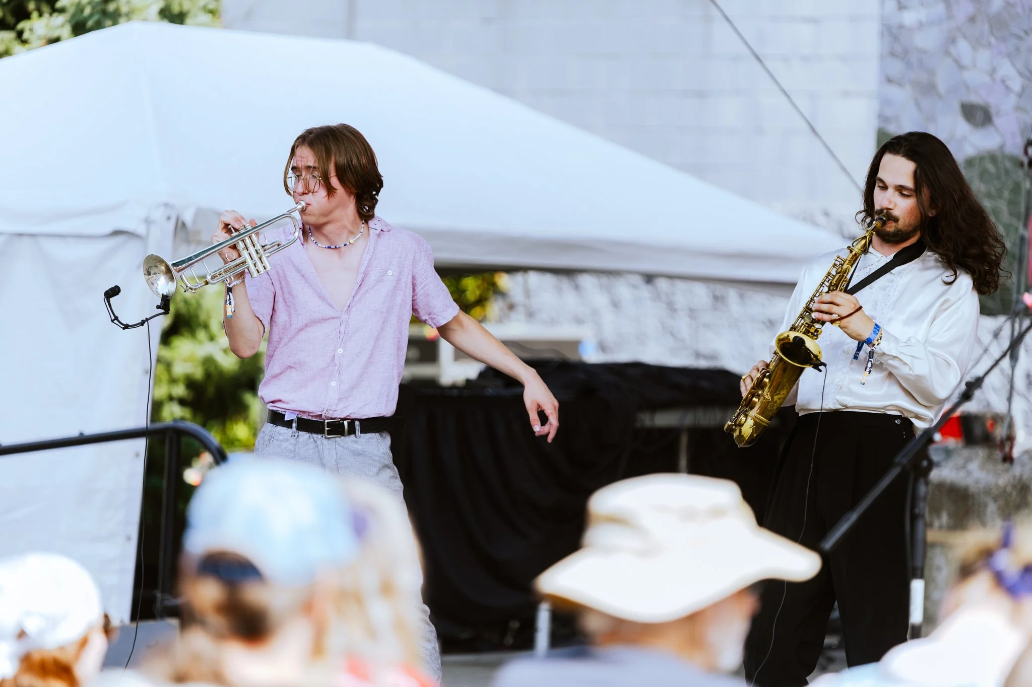 Two musicians perform at an outdoor event, one playing the trumpet and the other playing the saxophone, with an audience watching.