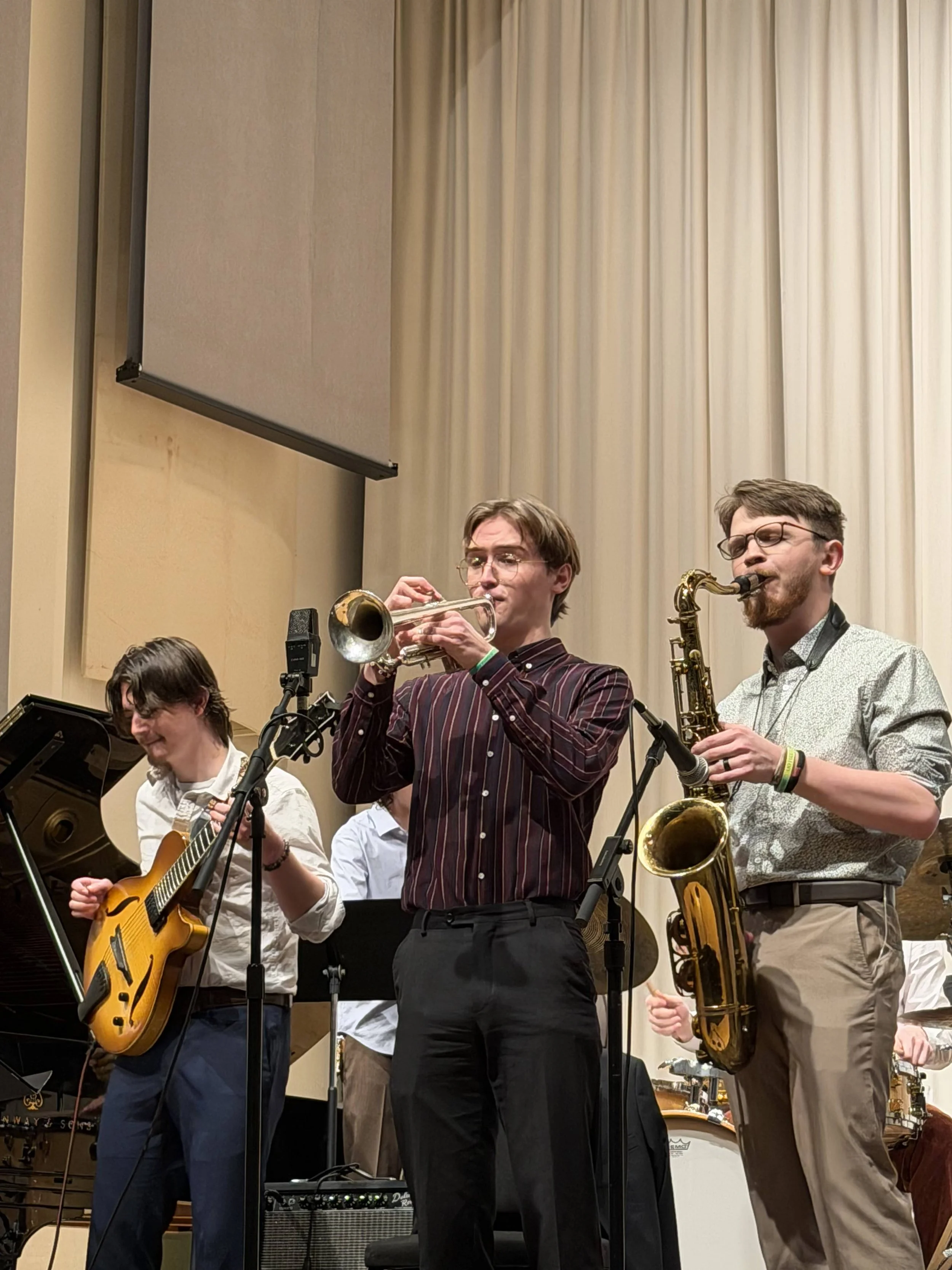 Three musicians playing on stage: a guitarist on the left, a trumpet player in the middle, and a saxophonist on the right.