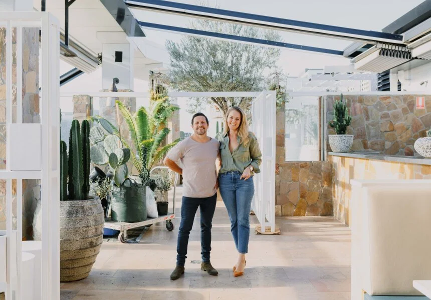 A man and woman standing inside a bright, modern indoor space with large windows and glass ceiling, surrounded by potted plants including cacti and succulents, smiling at the camera.