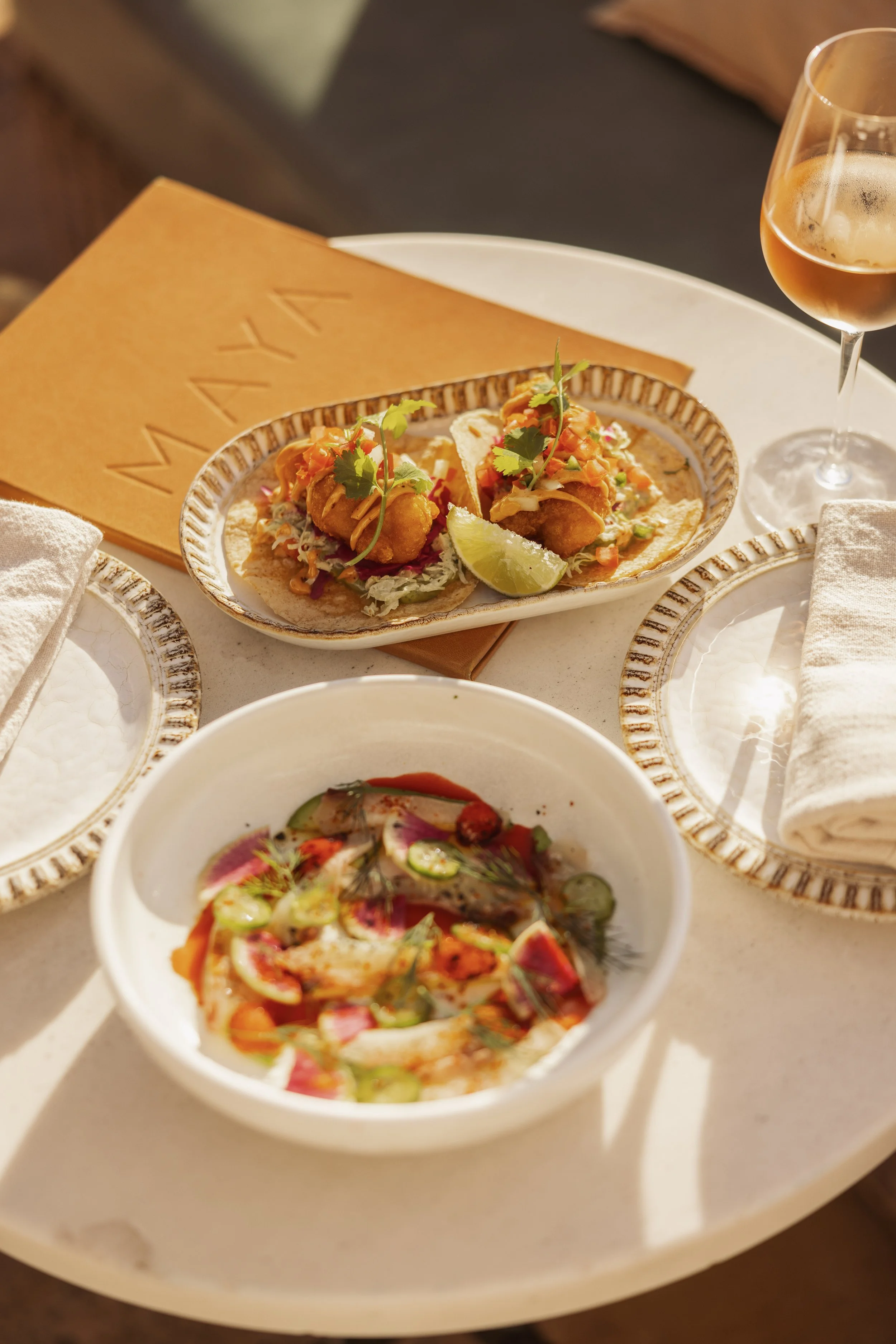 Tacos with fried fish, garnished with cilantro and lime, on a ceramic plate, with a bowl of ceviche and a glass of rosé wine on a white tray with napkins.