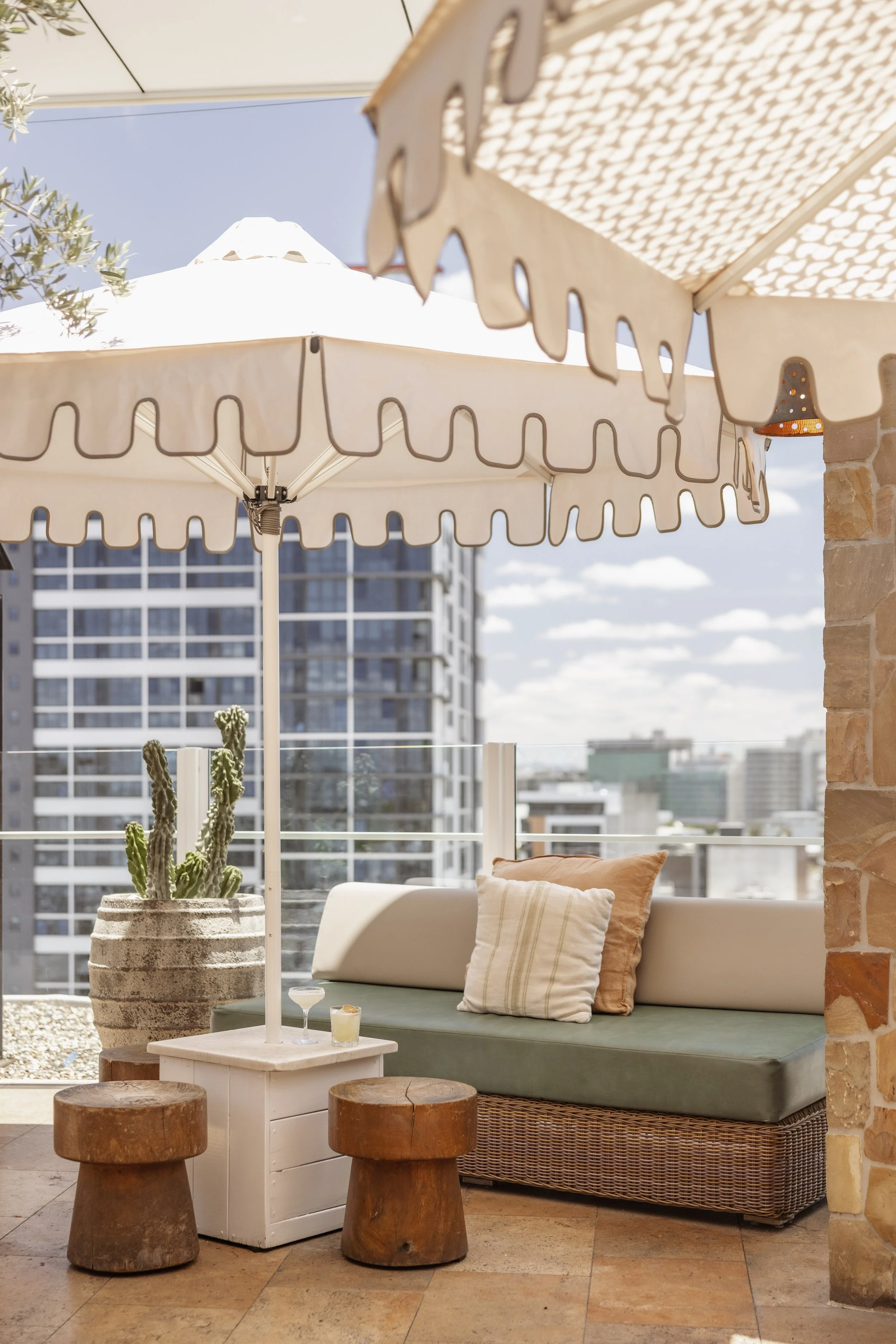 A sunlit rooftop patio with a large white umbrella, a wicker bench with cushions, a cactus in a pot, two wooden stools, and a small white table with drinks, overlooking a cityscape with tall buildings.