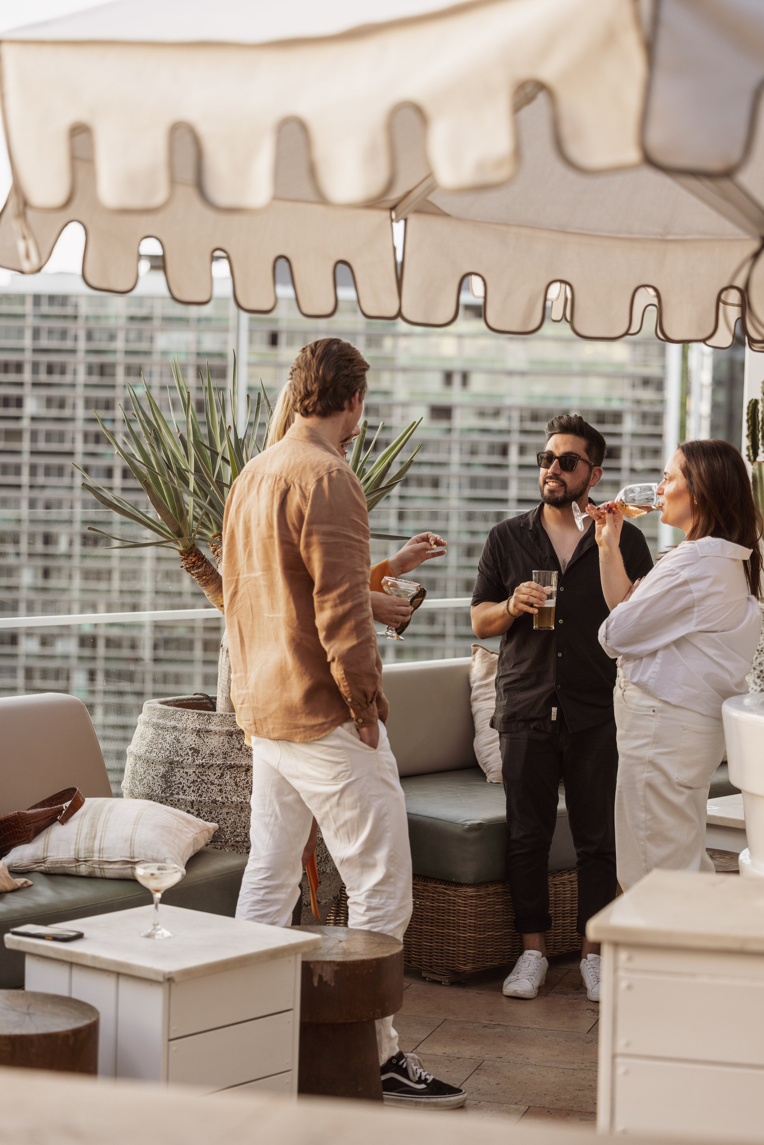 Four friends socializing on a balcony terrace with modern city buildings in the background, enjoying drinks and chatting under a large striped canopy.