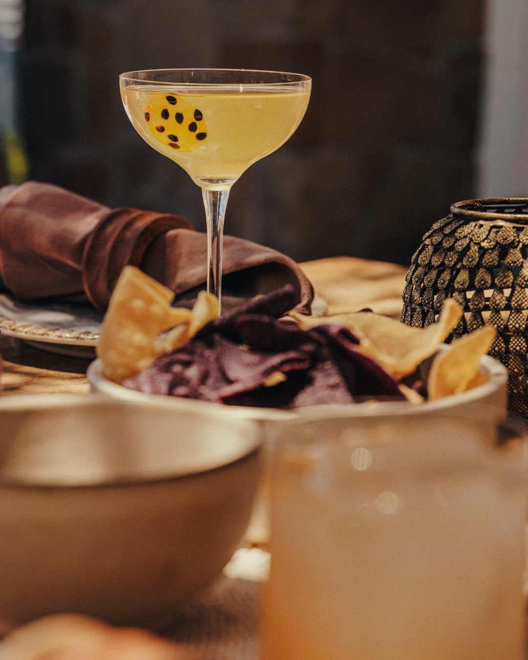 A cocktail glass with a yellow drink and a passion fruit slice on the rim, placed on a table with plates of chips and a bowl of purple chips in the foreground, surrounded by a brown napkin and decorative lantern.
