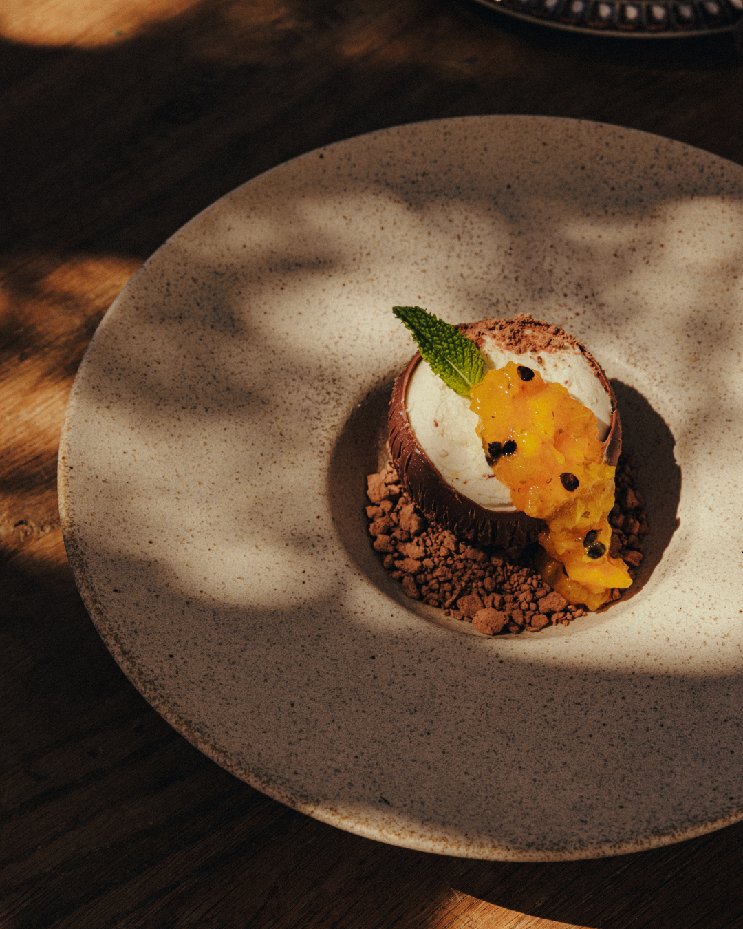 A dessert featuring a chocolate cup with a scoop of ice cream, topped with a yellow fruit compote, a mint leaf, and small black berries, served on a speckled beige plate with crushed chocolate and cocoa powder.