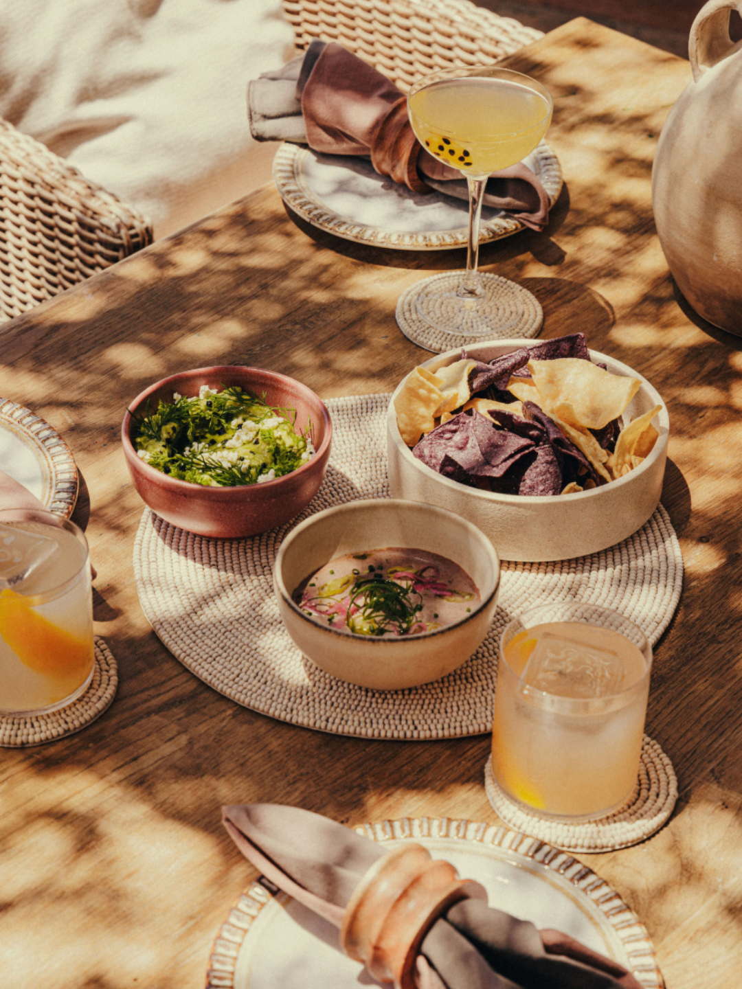 A rustic wooden table set with a salad, chips, dip, cocktails, and a napkin with utensils in an outdoor setting with dappled sunlight.
